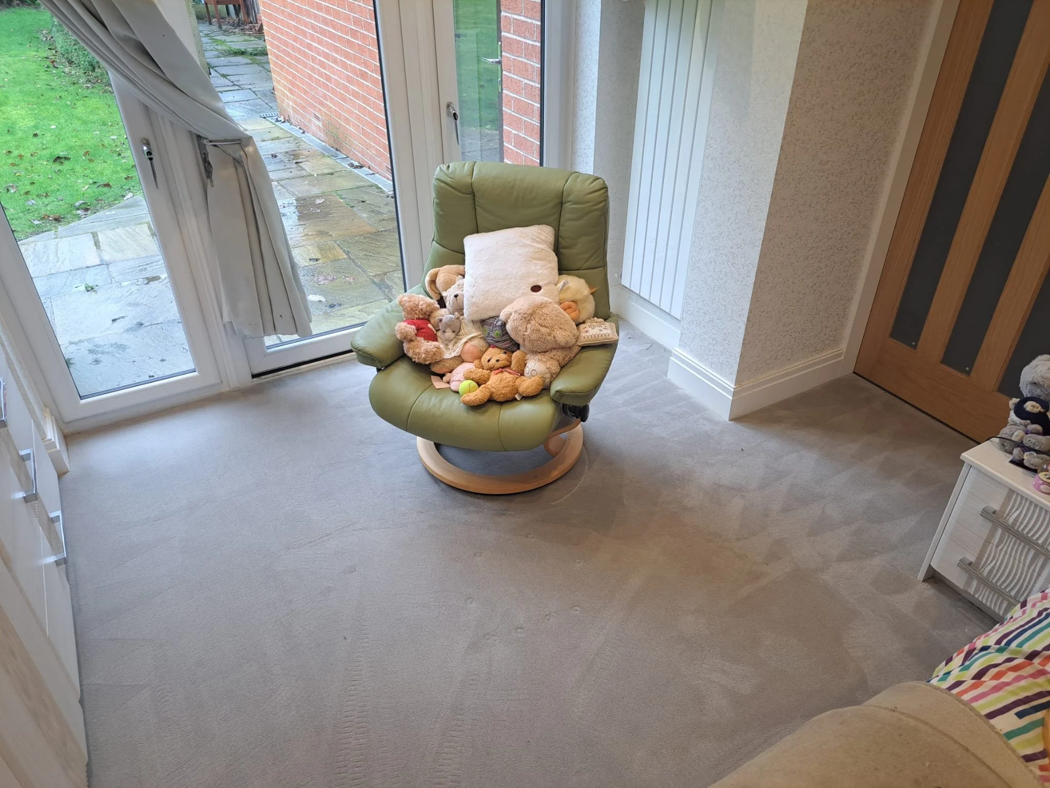 Indoor room with a pile of stuffed animals and pillows on a green rocking chair, next to large glass sliding doors leading outside to a wet patio and green yard.