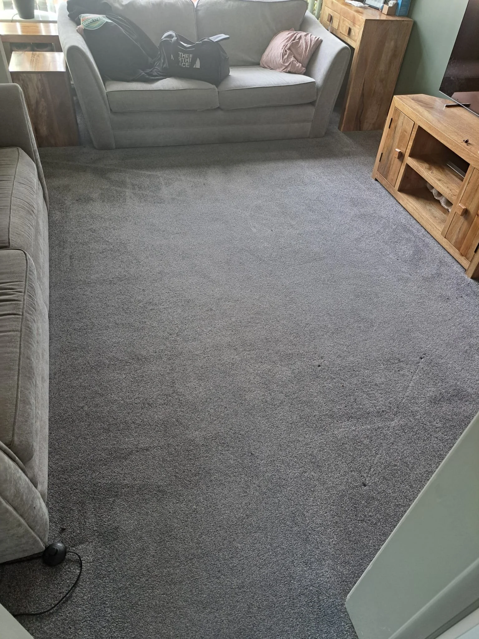 Living room with a gray sofa, a wooden cabinet, and a carpeted floor. Items on the sofa include a black bag, a pink pillow, and some clothes. There's a wireless speaker on the floor near the bottom left corner.