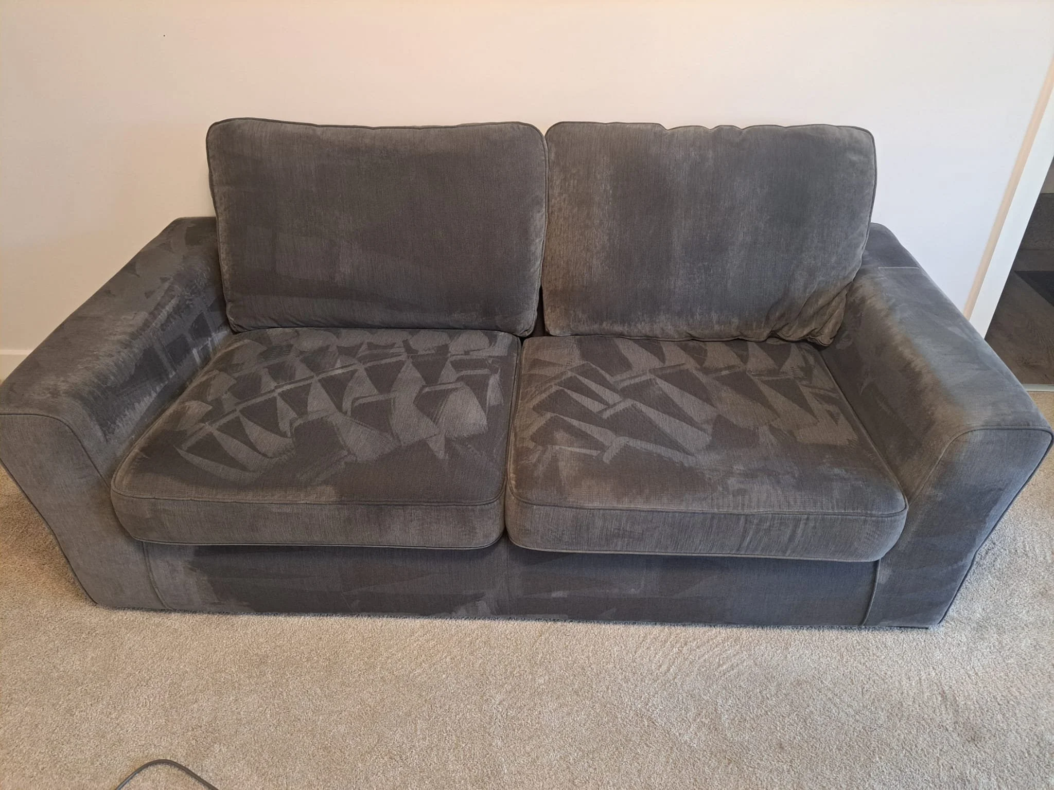 A gray upholstered couch with two large cushions, set against a plain white wall in a Room with beige carpet.
