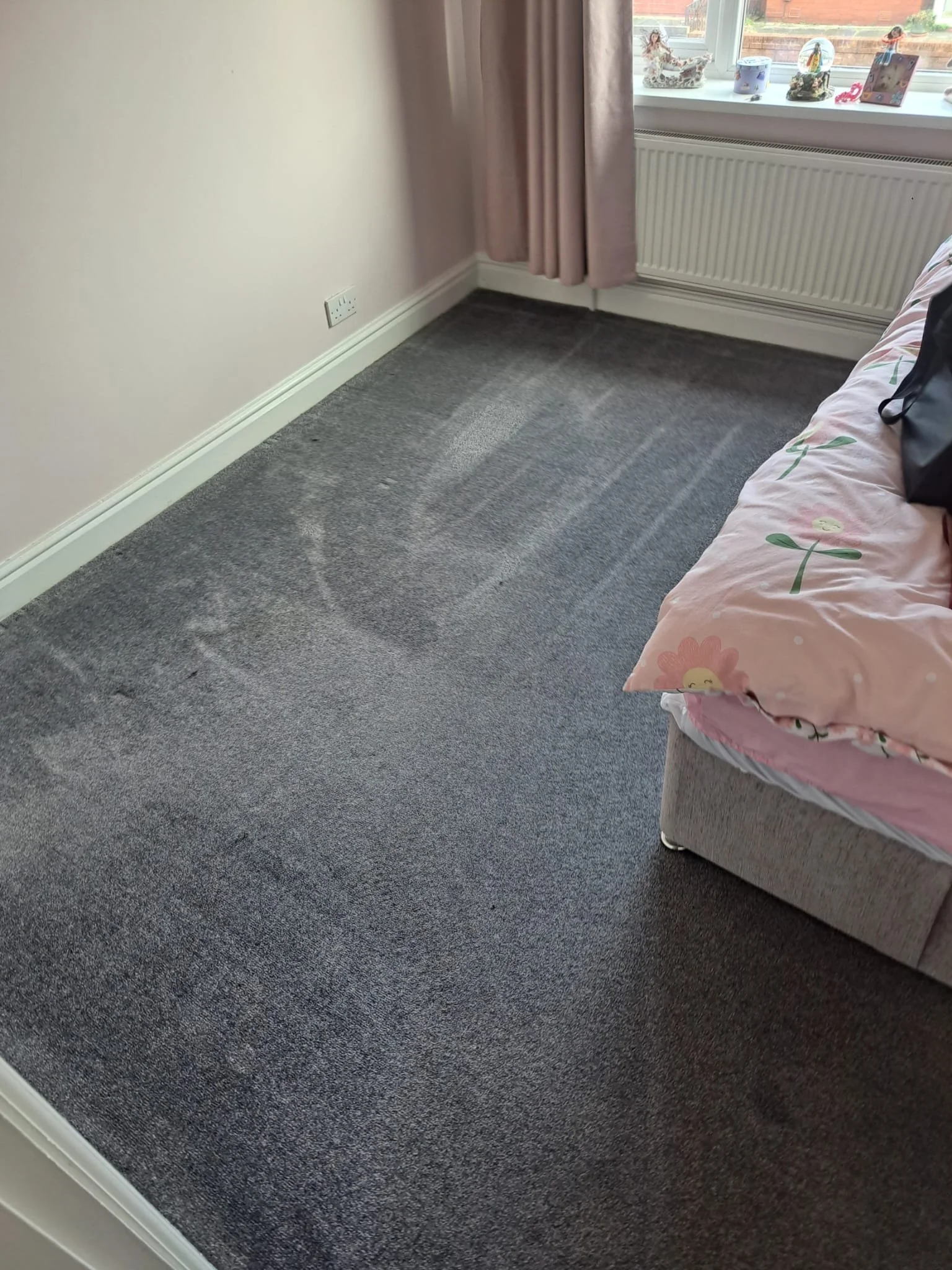 A corner of a room with grey carpet floor, white walls, pink curtains, and a window with decorative items on the sill. There is part of a couch with pink floral fabric and a black bag on it.