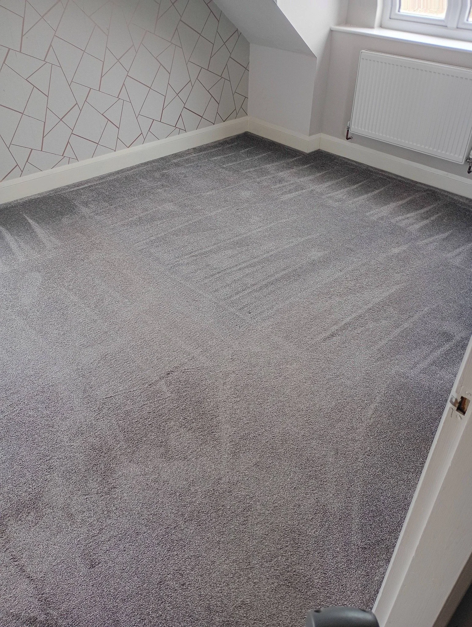 Empty room with grey carpet, patterned accent wall, white window, and radiator.