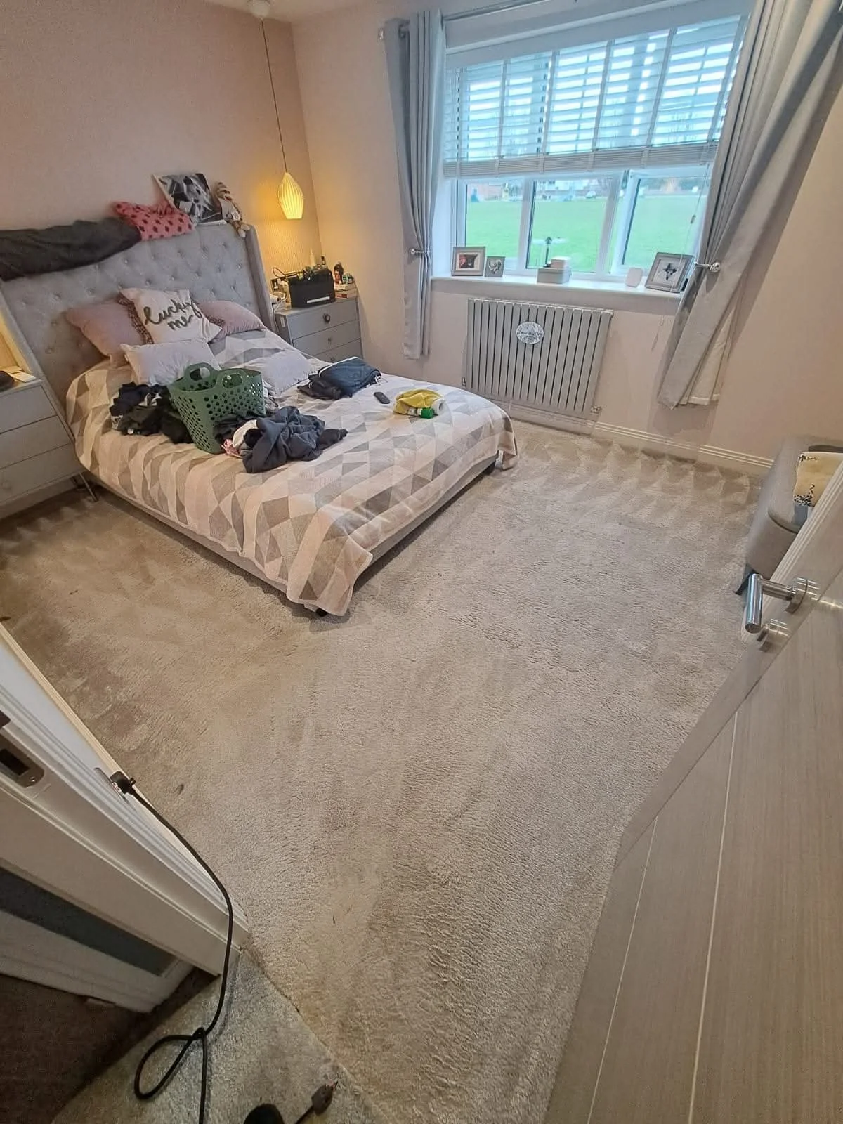 A bedroom with an unmade bed, clothing on top, and a green laundry basket. The room has a window with curtains, a beige carpet, and a nightstand with speakers and personal items. There are framed photos on the window sill and a view of green fields o