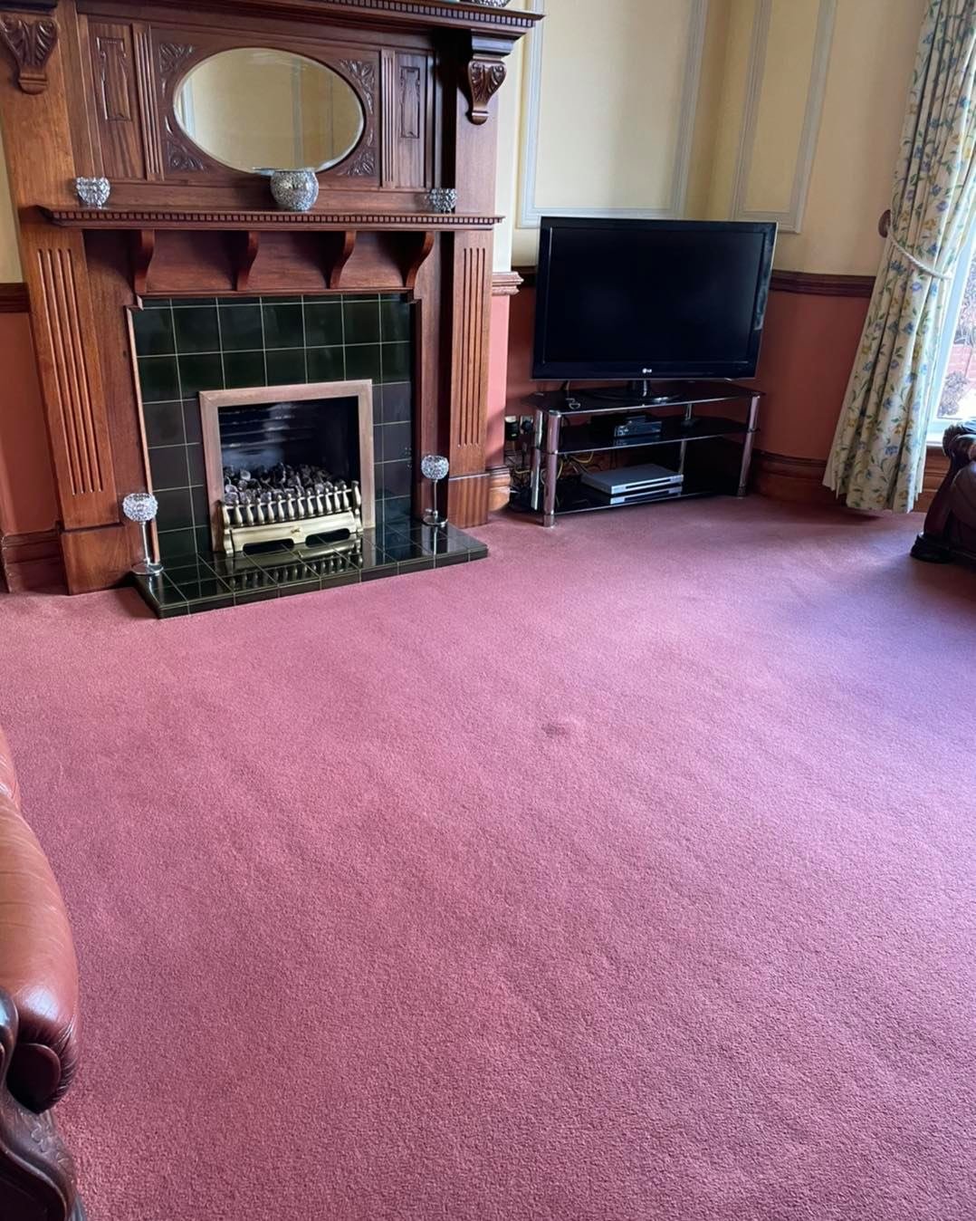 A living room with a pink carpet, a wooden fireplace with decorative glass ornaments, a flat-screen TV on a black stand, and a window with floral curtains.