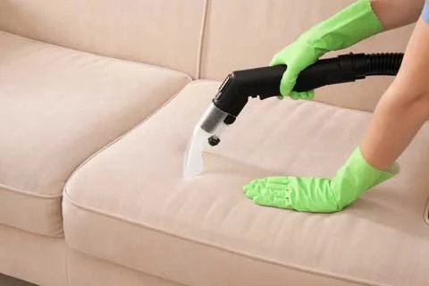 Person cleaning beige sofa with a handheld vacuum cleaner while wearing green rubber gloves.