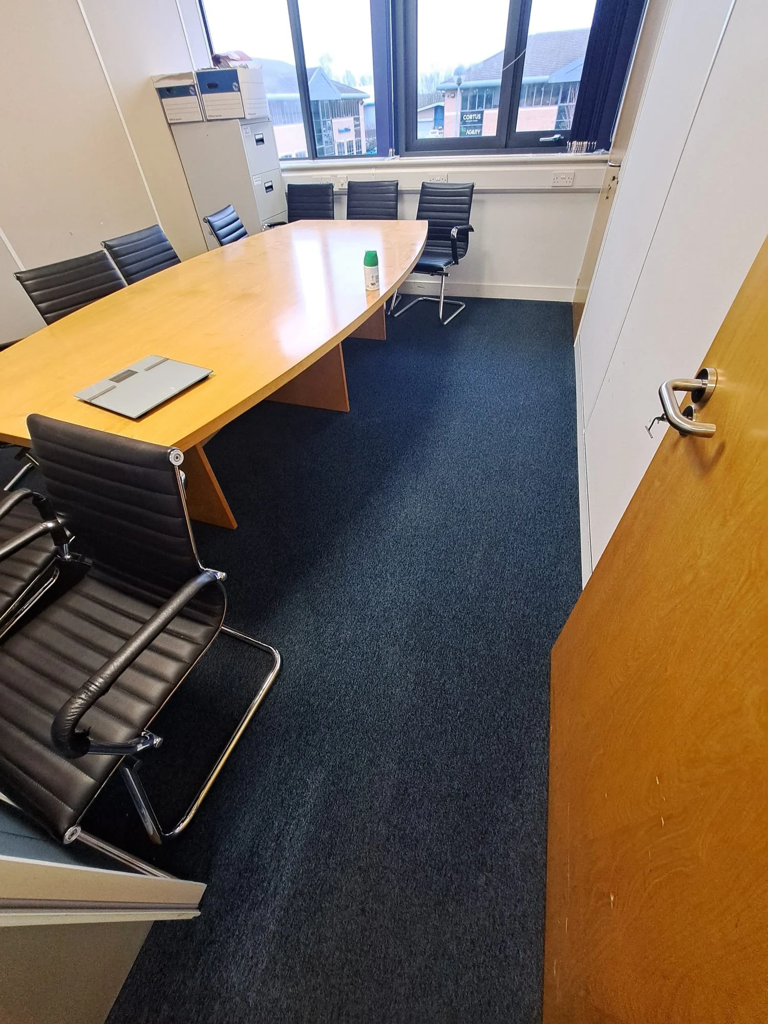 Empty office conference room with a wooden table, black chairs, a laptop, and a small container on the table. Large window with a cityscape view and filing cabinets against the wall.