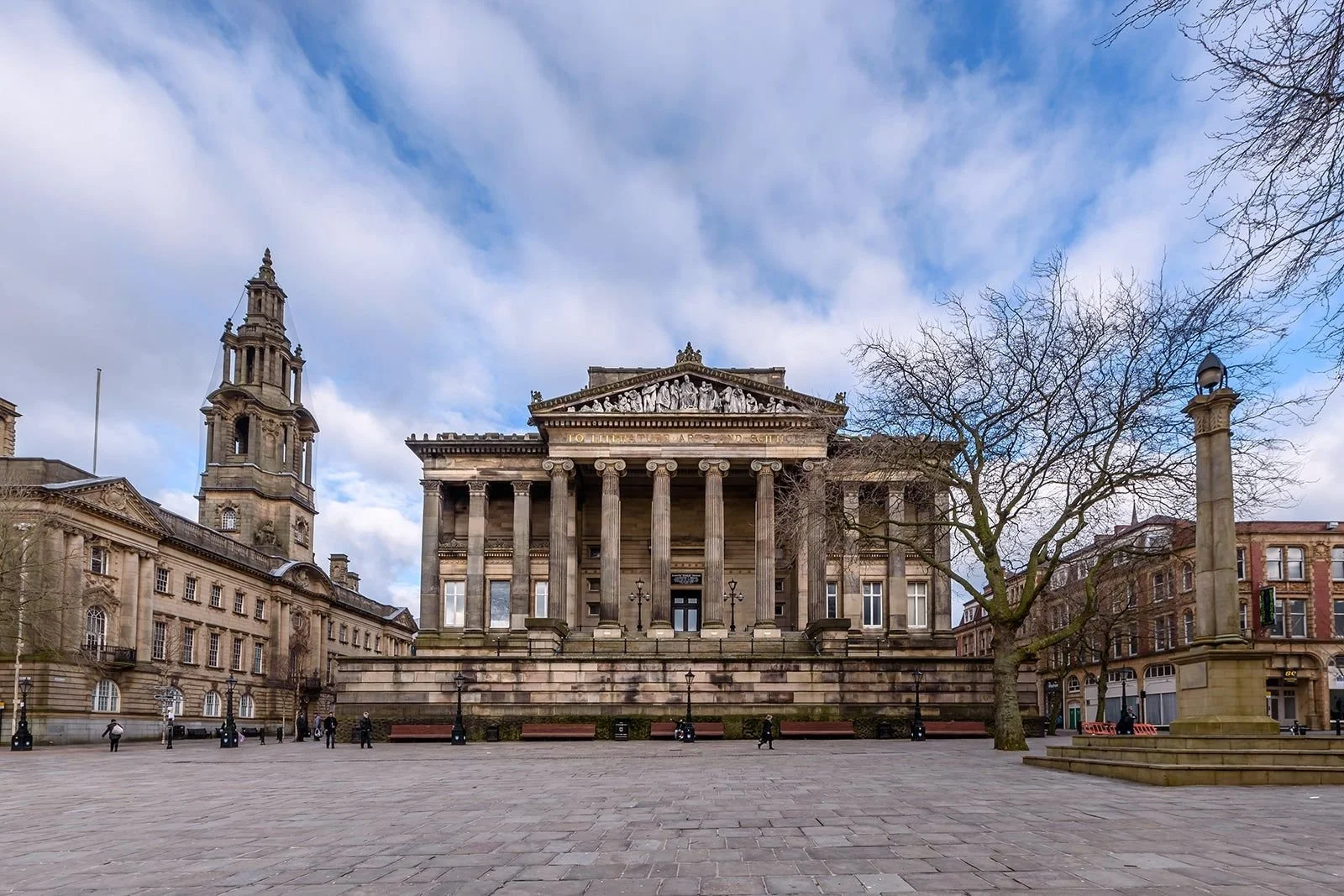 A historic stone building with columns and a triangular pediment, featuring sculptures and an inscription, situated in a town square with benches, a leafless tree, and a tall pillar, under a partly cloudy sky.