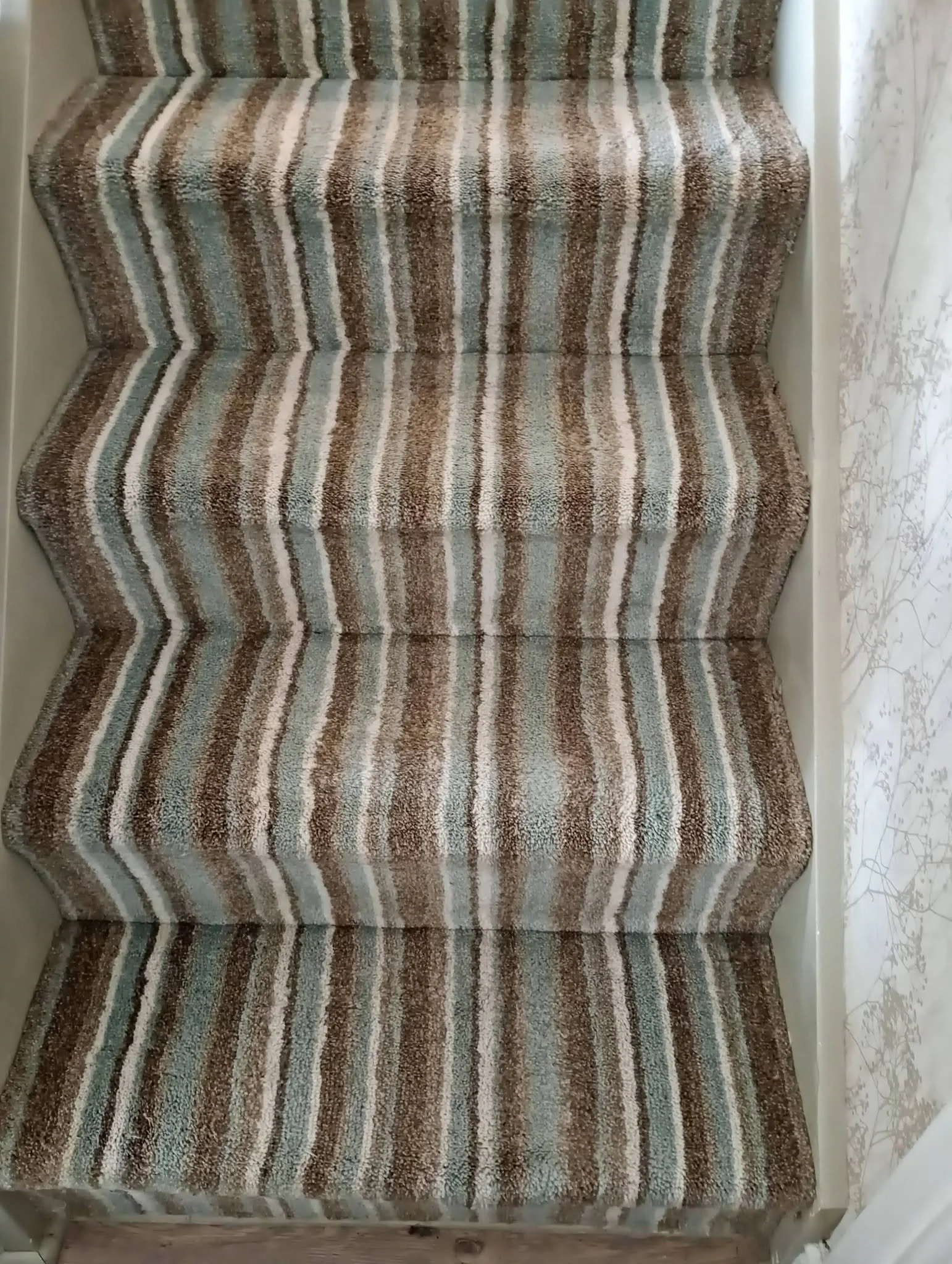 A staircase with striped carpet in shades of brown, beige, blue, and gray, with a marble wall on the right side and a white wall on the left side.
