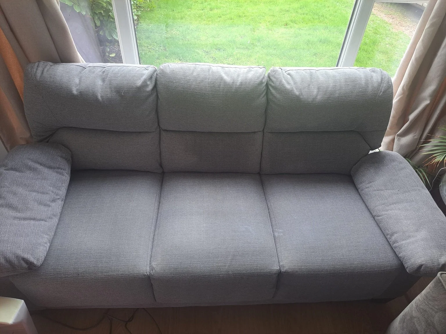 A gray fabric sofa with three back cushions, three seat cushions, and two armrests is positioned in front of a large window showcasing a grassy yard outside.