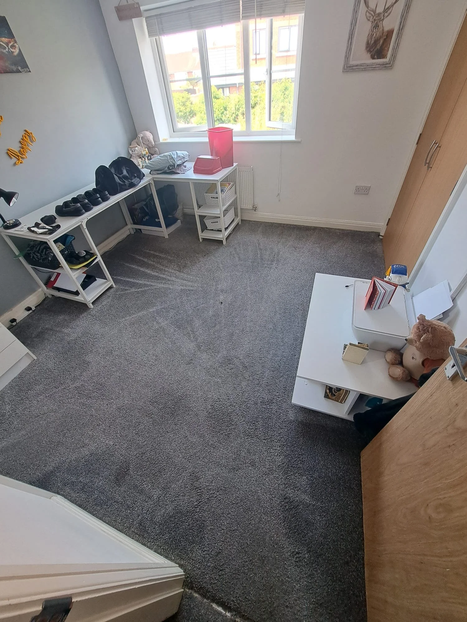 A room with a large window, gray carpet, white furniture including a shoe rack with shoes, a small white table with a few items, a wardrobe, and a desk with stuffed animals and papers.