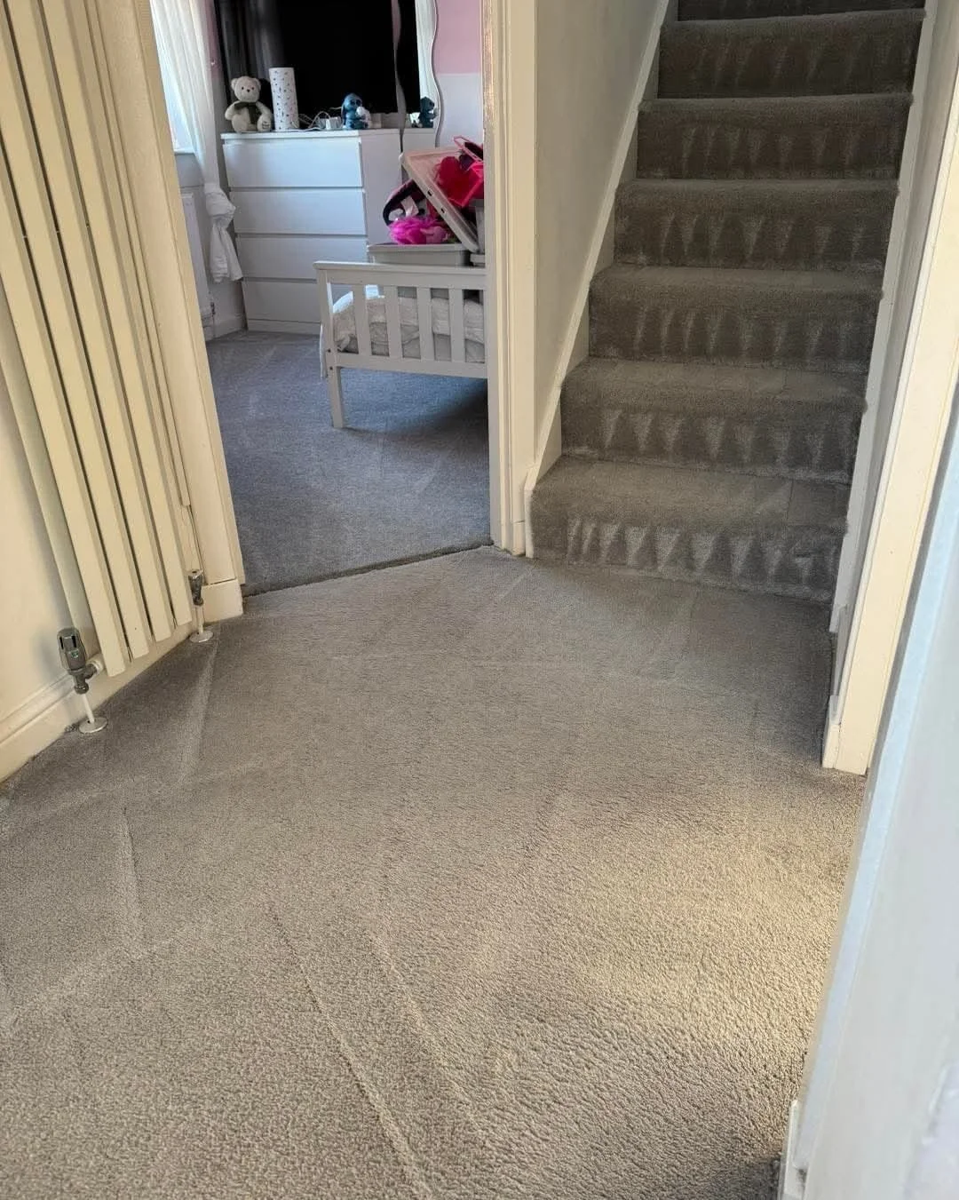 View of an entryway with beige carpet, a radiator on the left, leading into a bedroom with grey carpet, a white crib, and a window with toys on the sill. A staircase with grey carpet is on the right.