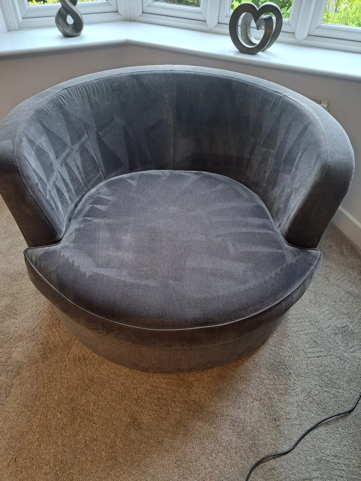 A round, gray upholstered armchair with a curved backrest, placed on a beige carpet in front of a bay window with decorative metal hearts on the window sill.