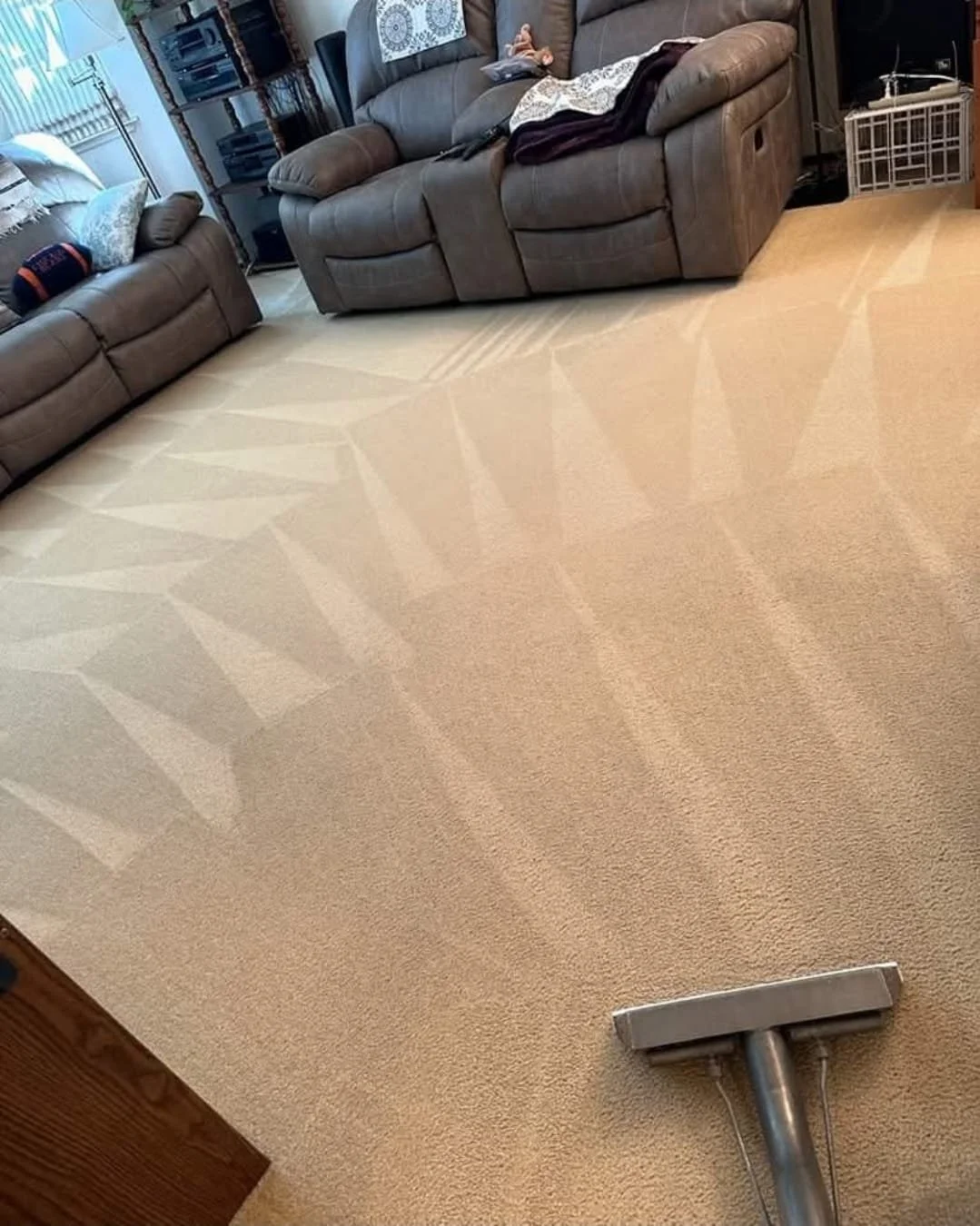 A person vacuuming a beige carpet in a living room with two brown sofas, a window with blinds, and a metal entertainment center.