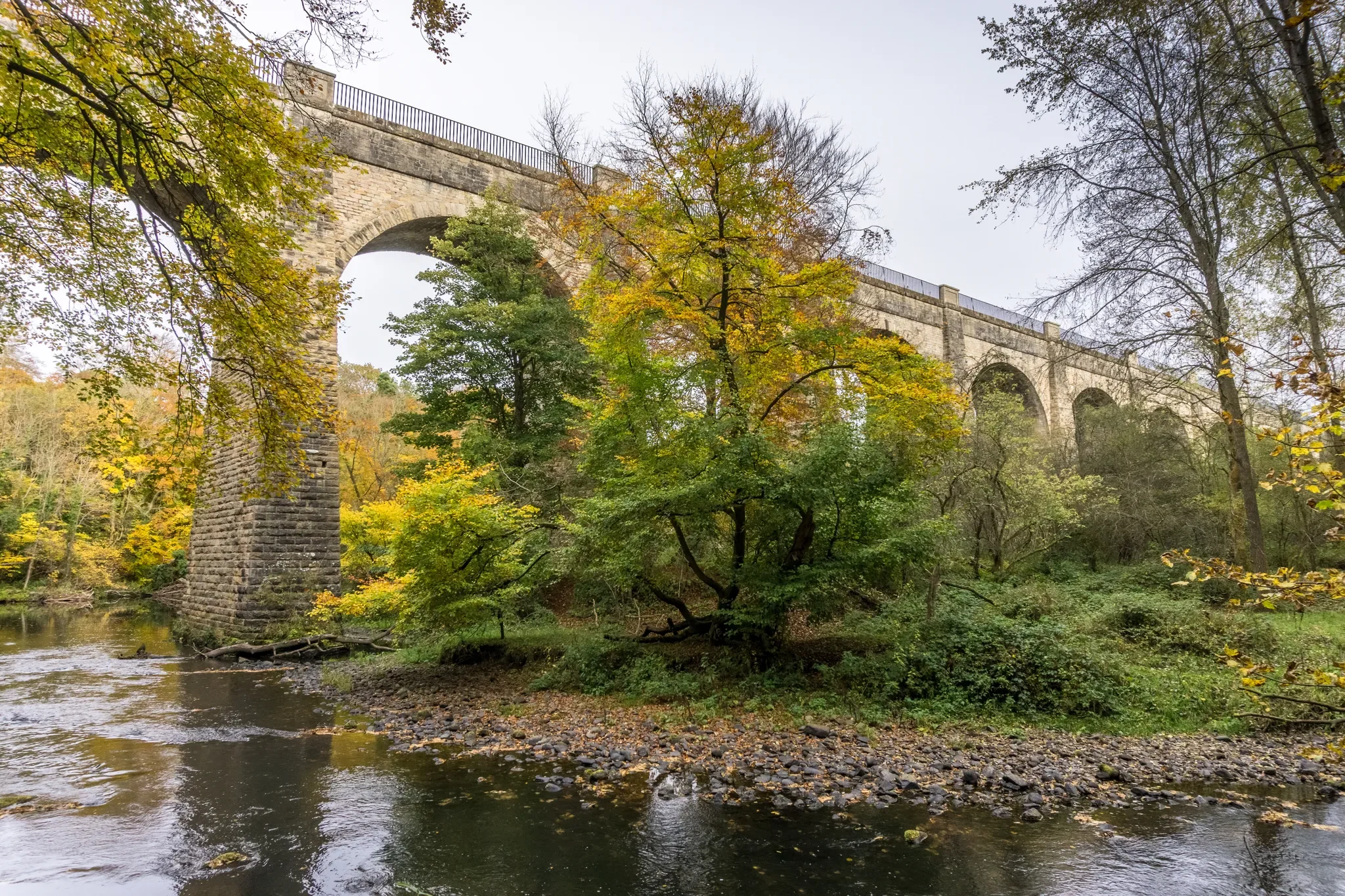 A scenic view of a stone railway bridge over a river with trees displaying fall foliage surrounding the area.