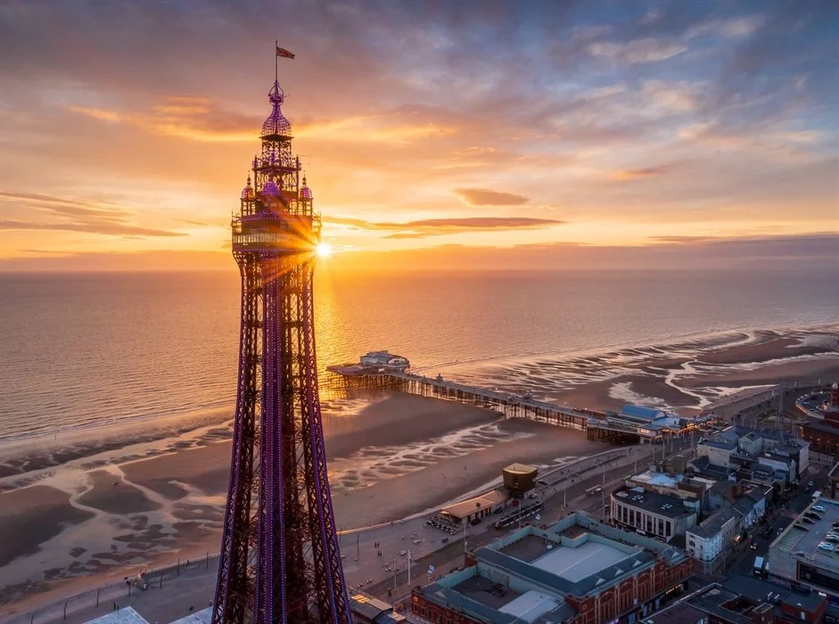 Sunset over a pier at the beach with an illuminated tall tower, possibly the Blackpool Tower, in the foreground.