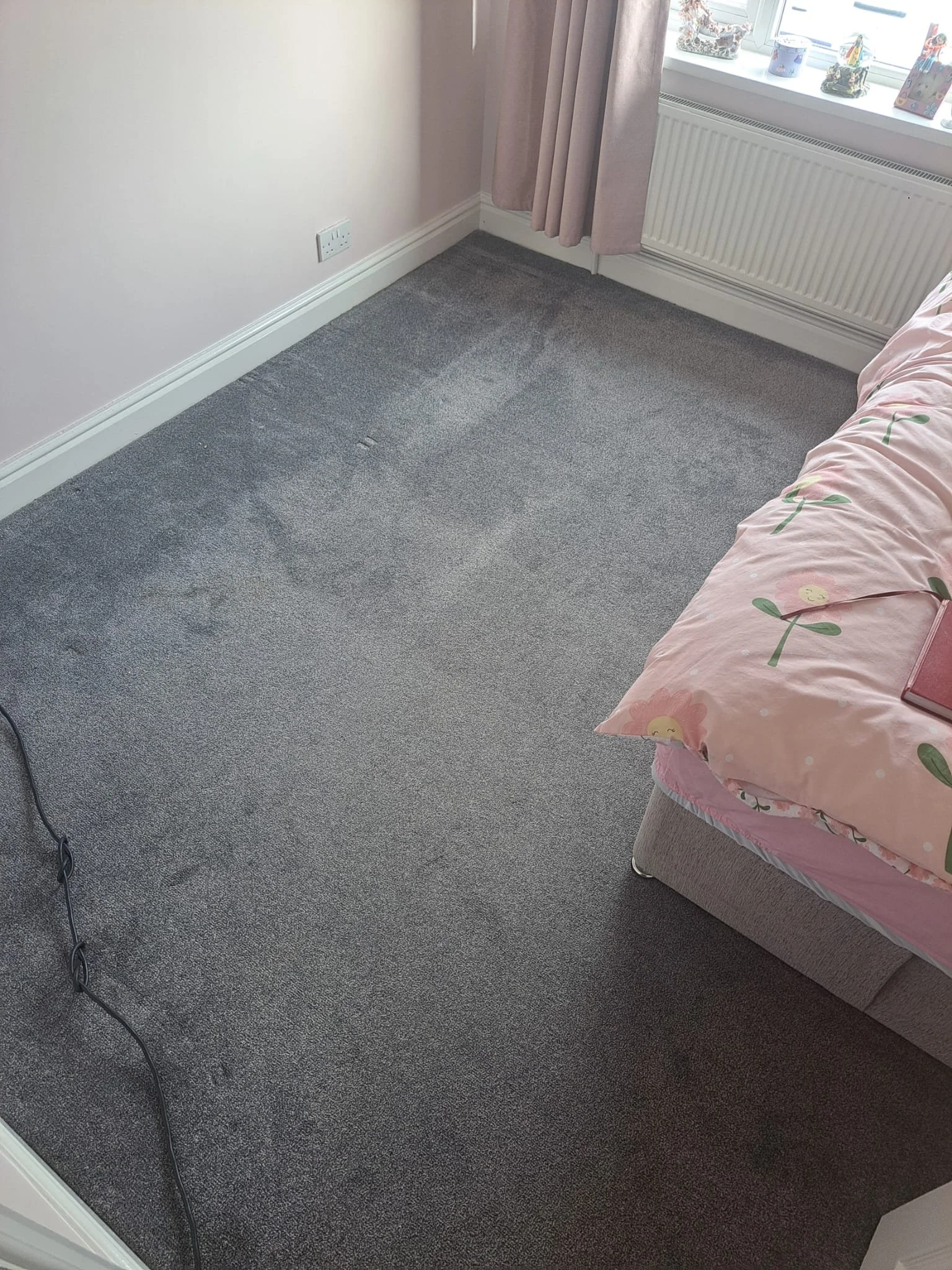 A corner of a bedroom with pink walls, carpeted floor, pink curtains, a bed with pink floral bedding, and decorations on the windowsill.
