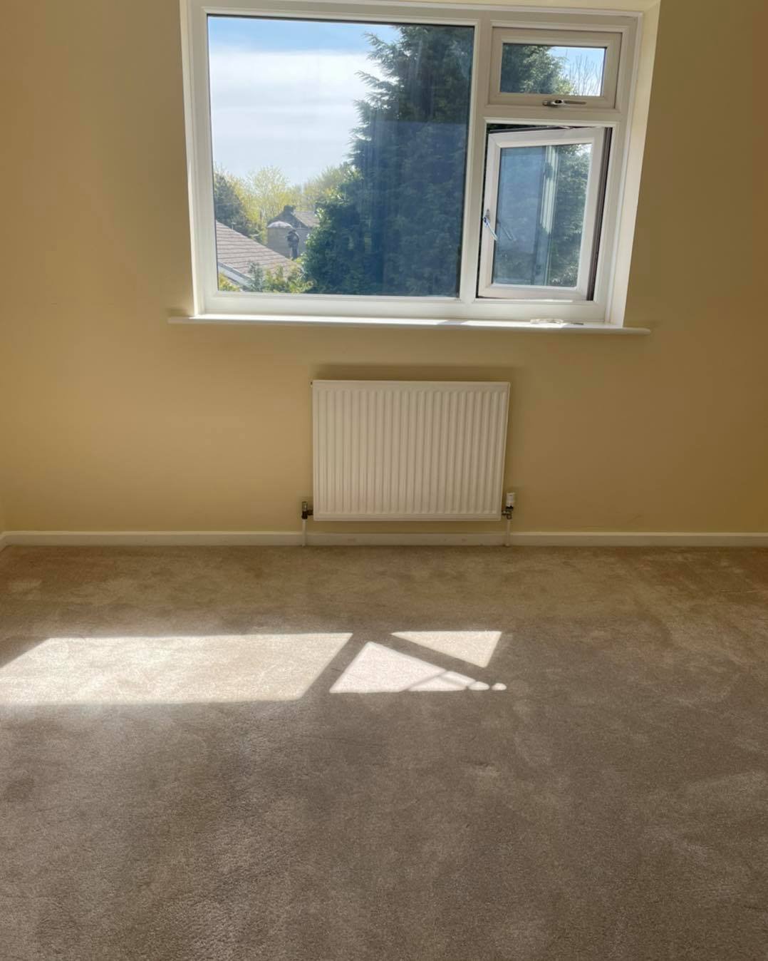 An empty room with beige walls, a window, and a white radiator. Sunlight streams through the window, casting a shadow on the light-colored carpeted floor.