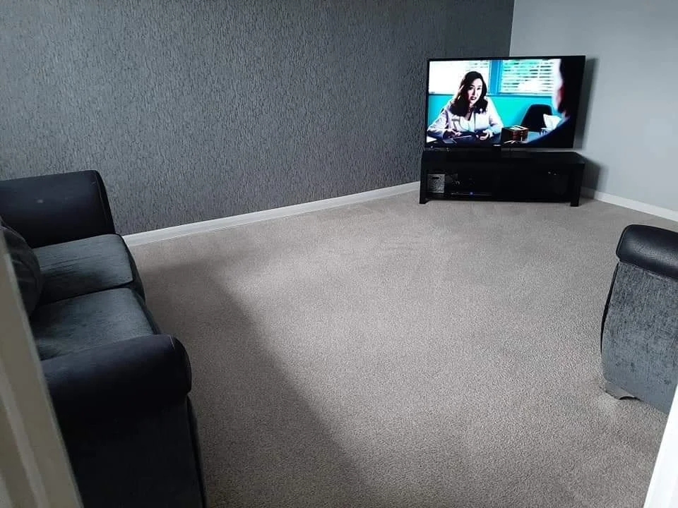 Empty living room with gray walls, beige carpet, black sofa, gray upholstered chair, and a flat-screen TV on a stand showing a woman talking.