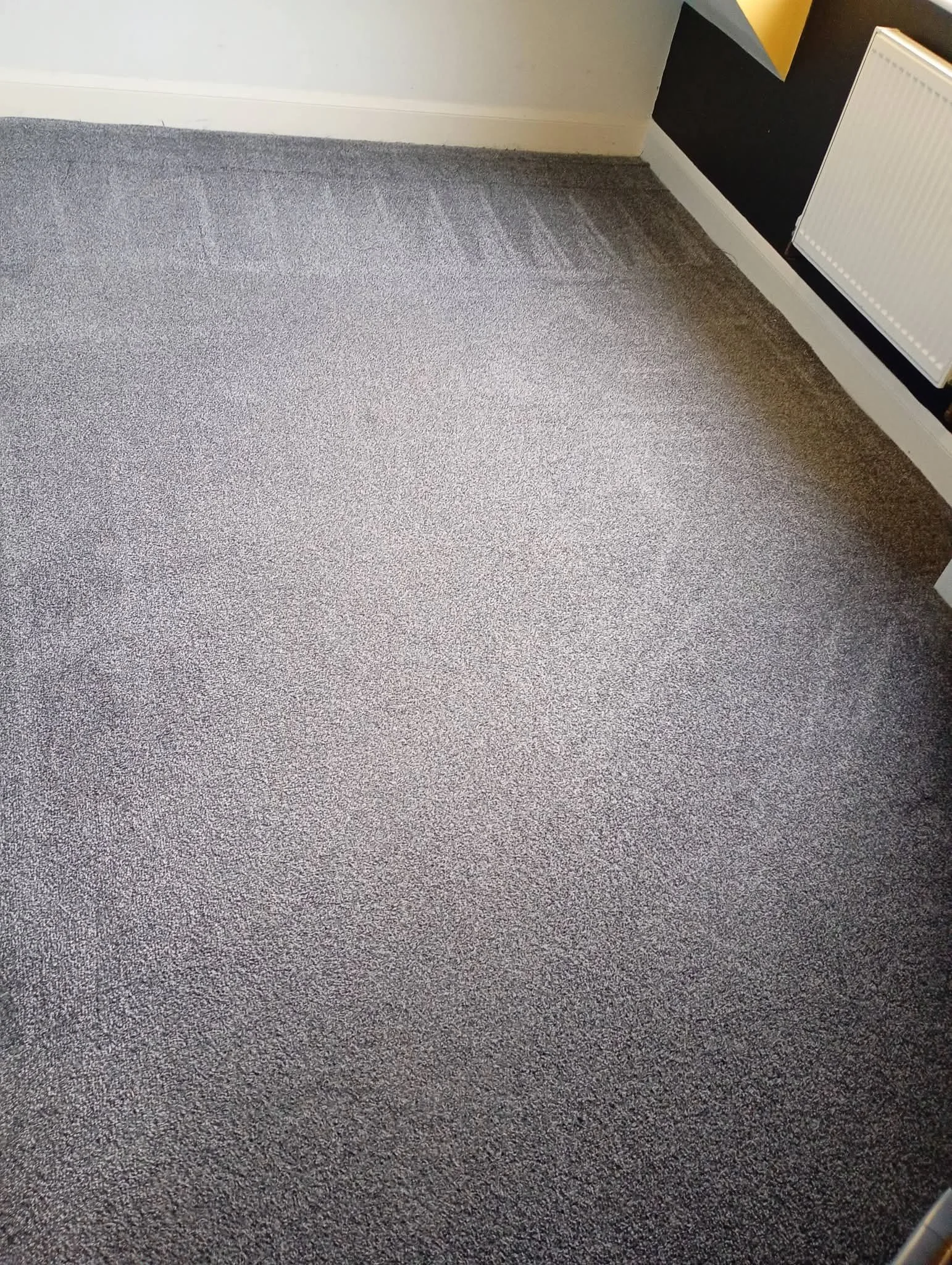 A gray textured carpet floor corner with a white baseboard, a white wall, a black wall with posters, and a white radiator.