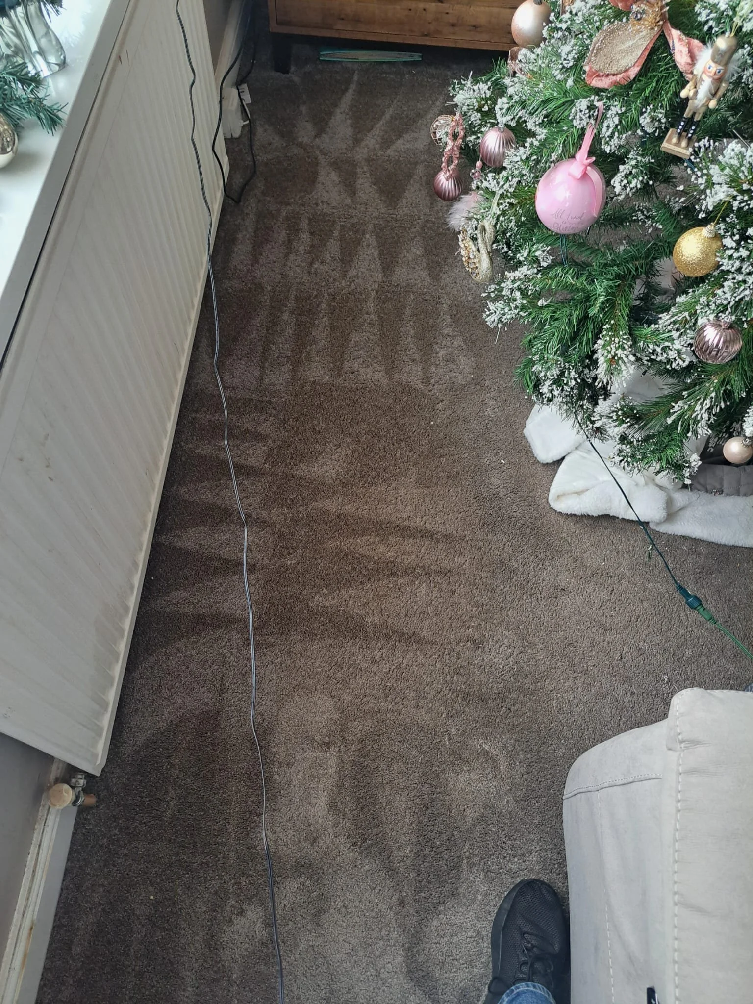 A decorated Christmas tree with pink, gold, and white ornaments, located next to a white cushioned chair. There is a white fluffy tree skirt underneath. A black electrical cord runs along the carpeted floor, next to a white radiator and a small woode