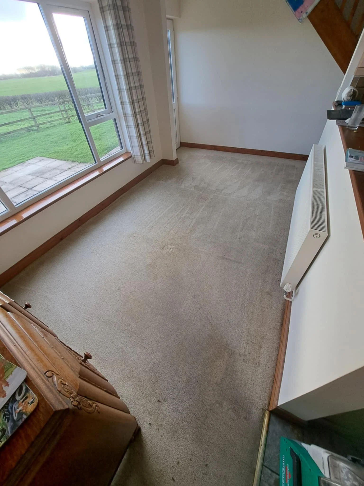 Empty room with large windows looking out onto a green field, beige carpet, white walls, and wooden baseboards. Part of a wooden cabinet and scattered items are visible in the room.