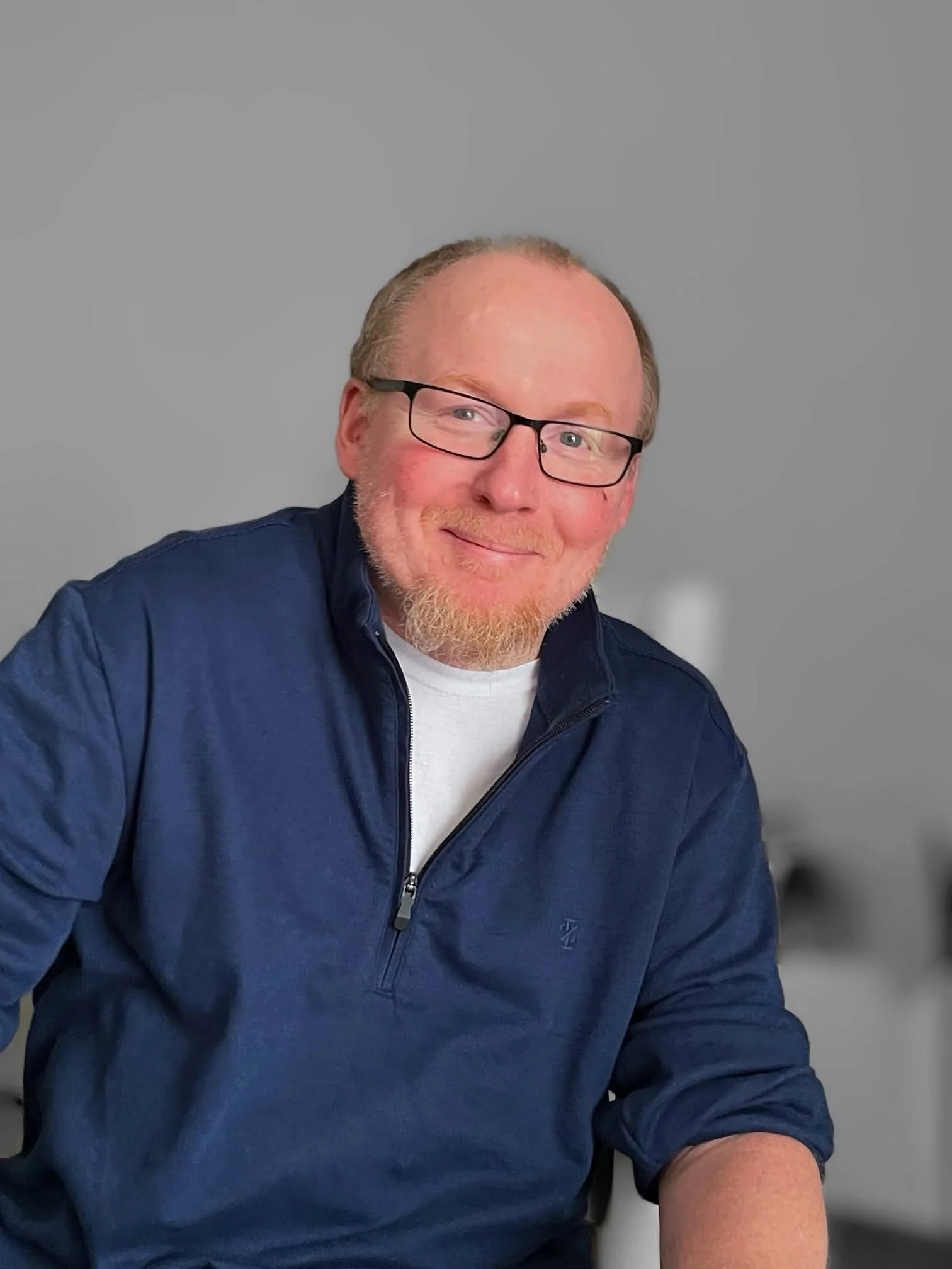 A smiling man with glasses, a beard, wearing a navy blue jacket over a white shirt, seated against a gray background.