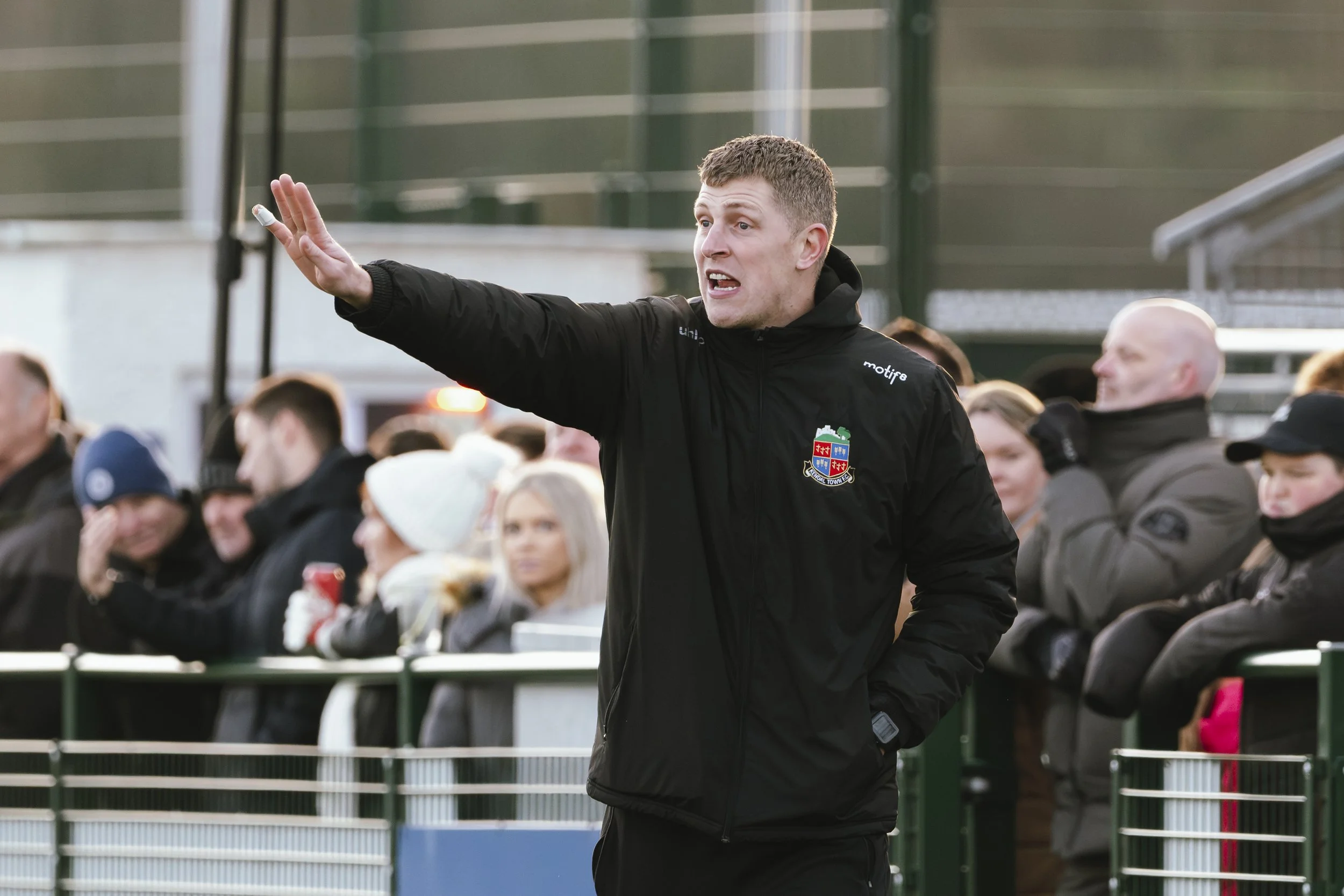 A man in a black jacket with a crest on it is gesturing with his right hand while speaking to a crowd behind a metal fence at an outdoor sports event.