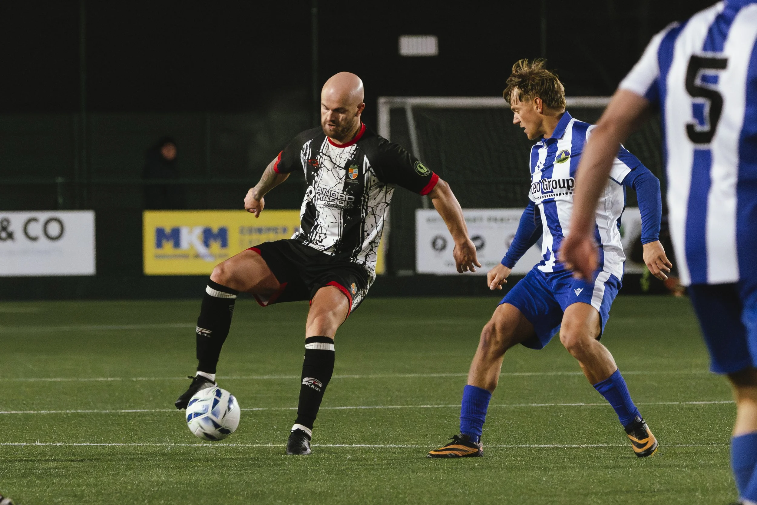 Soccer players in action on the field, one in black and white jersey about to kick the ball, others in blue and white jerseys attempting to block.
