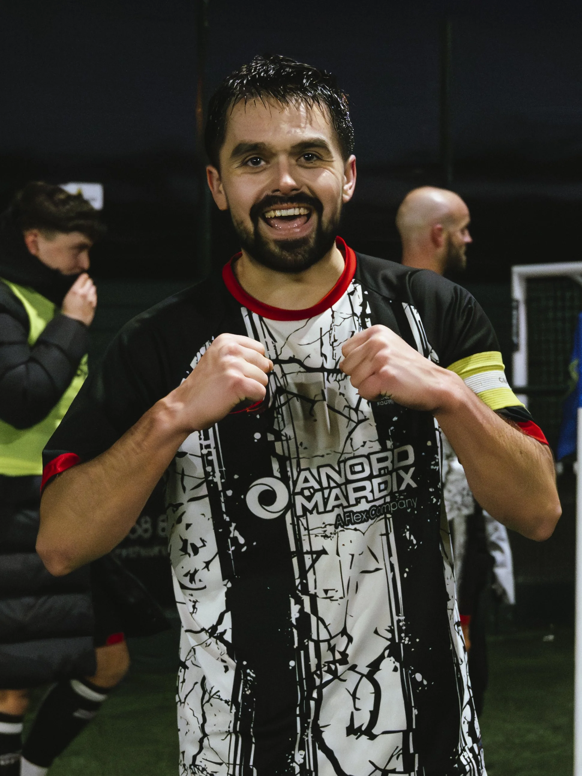A soccer player in a black and white jersey celebrating with clenched fists, smiling, on a field with other people in sports attire in the background.