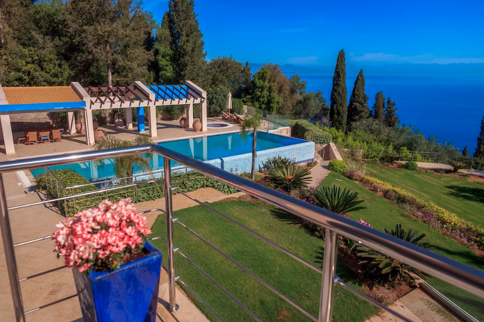 A luxurious outdoor pool area overlooking the ocean, with lounge chairs, shaded cabanas, and lush landscaping including palm trees and flowering plants.