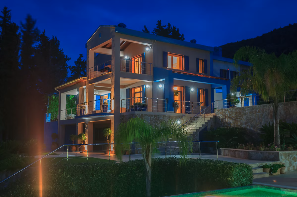 Modern multi-story house with large windows and balconies illuminated at night, surrounded by trees and landscaping.