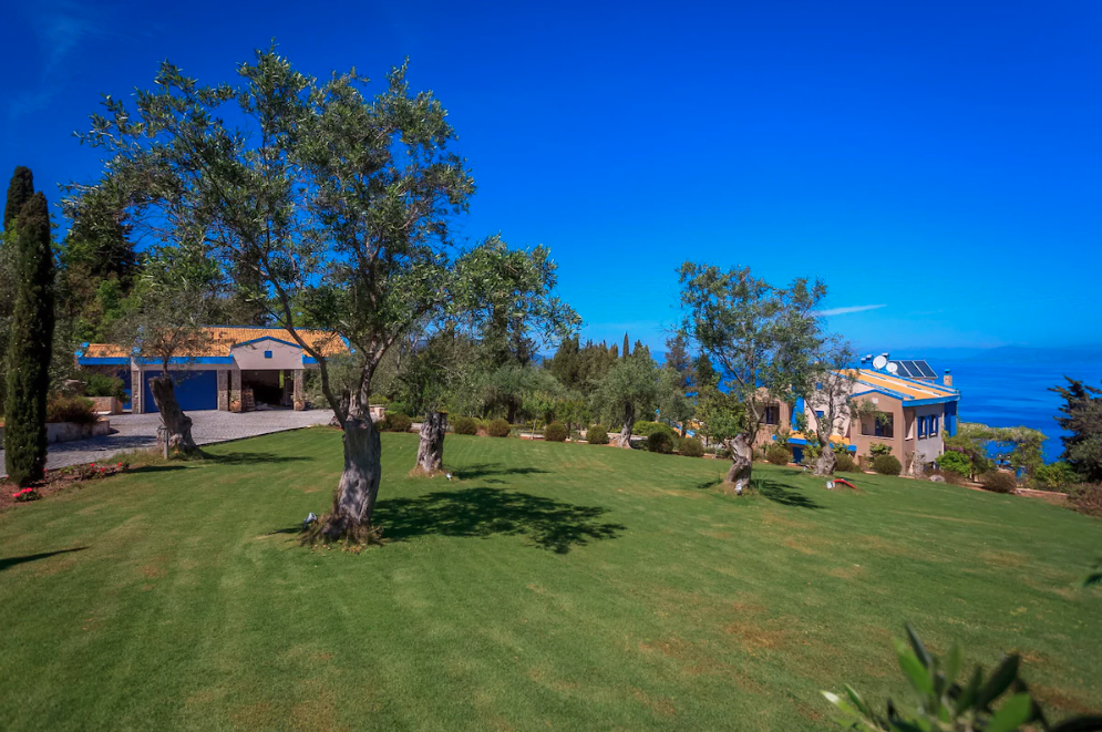 A large green lawn with trees, a house with solar panels on the roof, and a view of the ocean in the background.