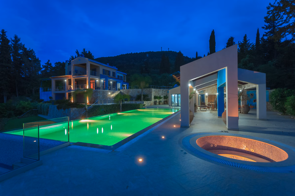 Luxury villa with illuminated swimming pool and hot tub at dusk, surrounded by hillside and trees.