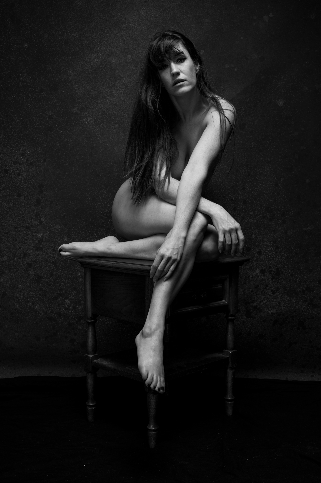 Schwarz-Weiß-Fotografie einer nackten Frau mit langen Haaren, die auf einem kleinen Holzstuhl sitzt, auf dem Kopf und einer Hand auf dem Knie, vor dunklem, rauh strukturieren Hintergrund.