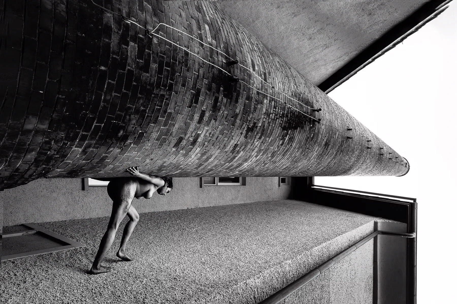 Schwarz-weiß-Foto eines nackten Mannes, der einen langen, zylindrischen, aus Ziegelsteinen gebauten Kamin auf einem betonierten Balkon hochklettert.