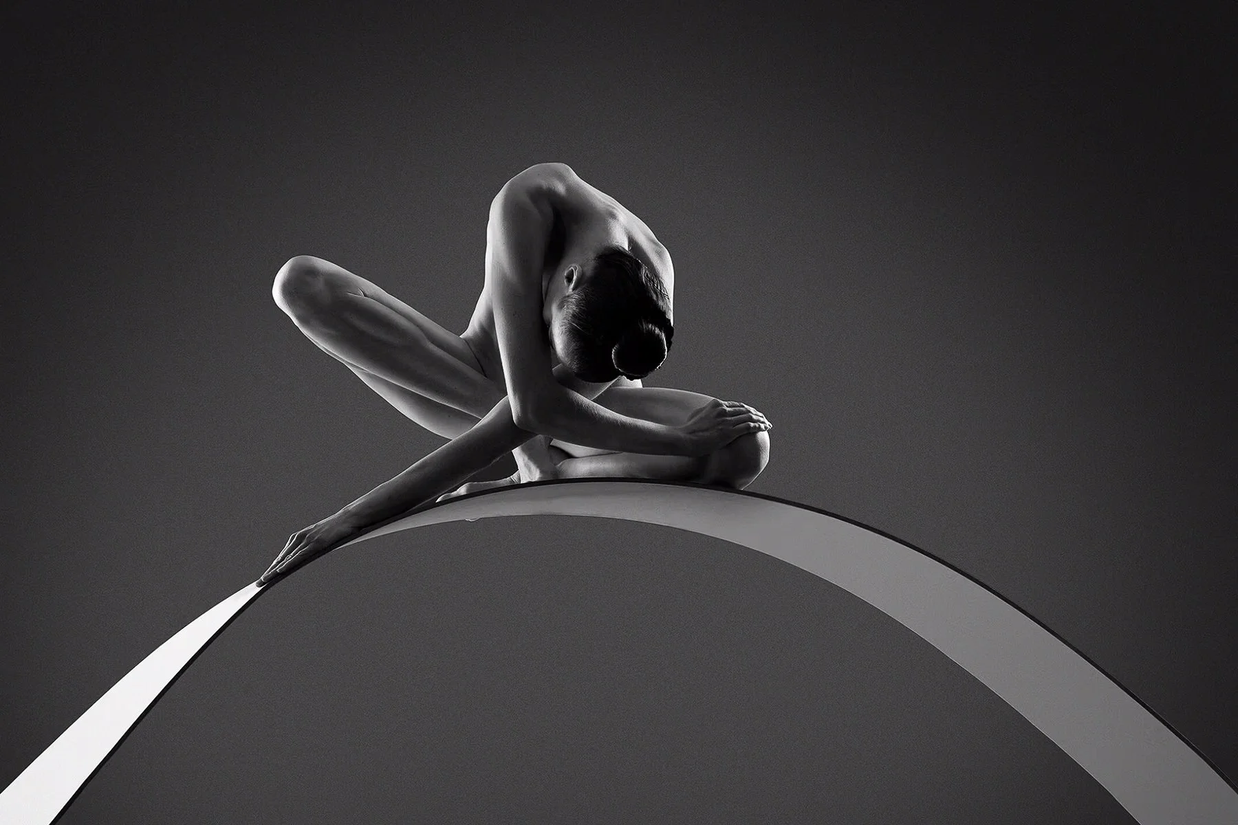 Schwarz-Weiß-Bild einer jungen, gestreiften Tänzerin, die auf einem weißen Bogen balanciert, in einer gekrümmten Haltung mit Blick nach unten.