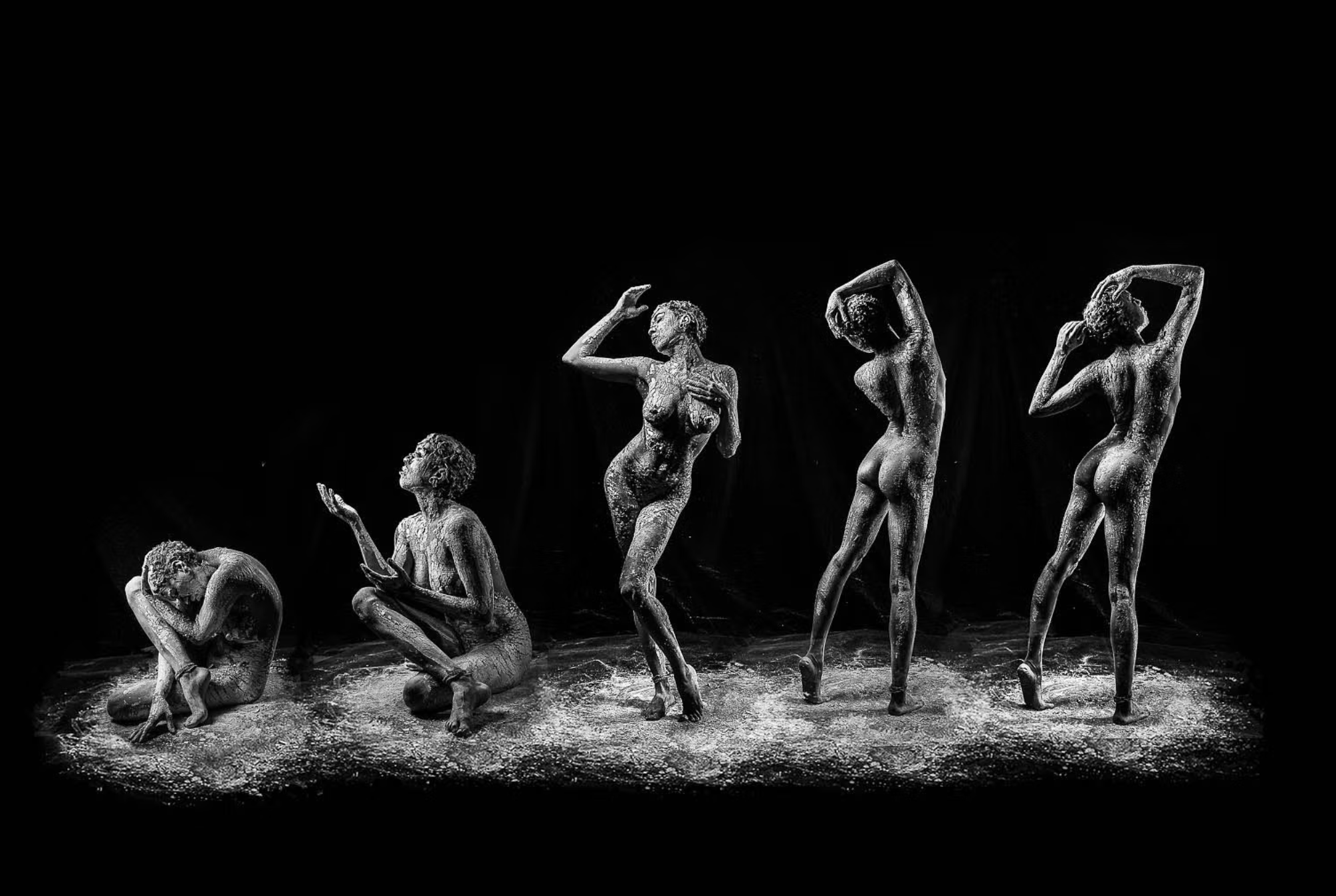 Schwarz-weiß Skulpturen von fünf weiblichen Figuren in verschiedenen Posen, auf einer dunklen Bühne mit hellem Staub oder Sand auf dem Boden