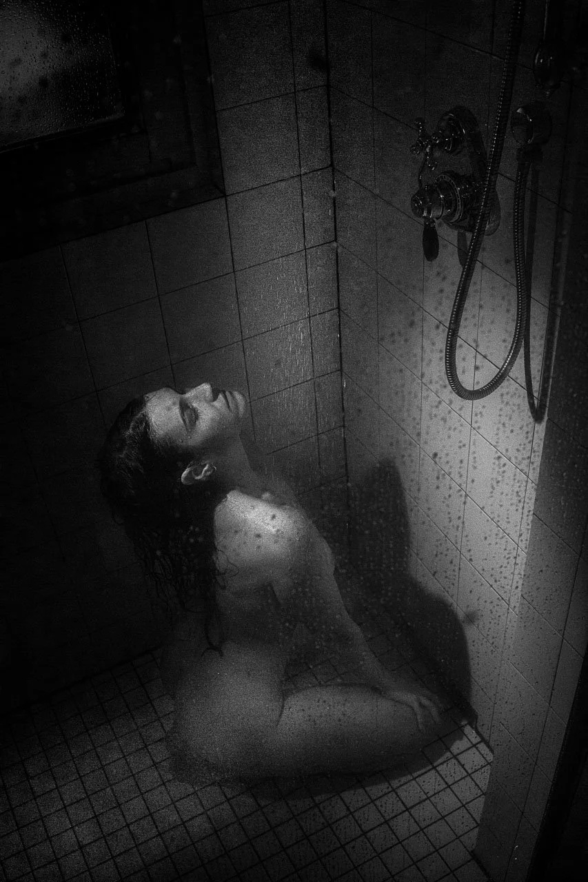 Nackt sitzende Frau im Duschraum, Blick nach oben, nasse Haare, Wasser auf der Haut, Duschkopf und Wasserstrahlen sichtbar.