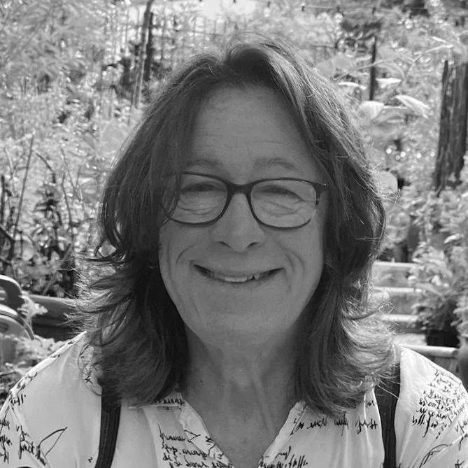 A smiling woman with shoulder-length hair and glasses, outdoors surrounded by trees and plants.