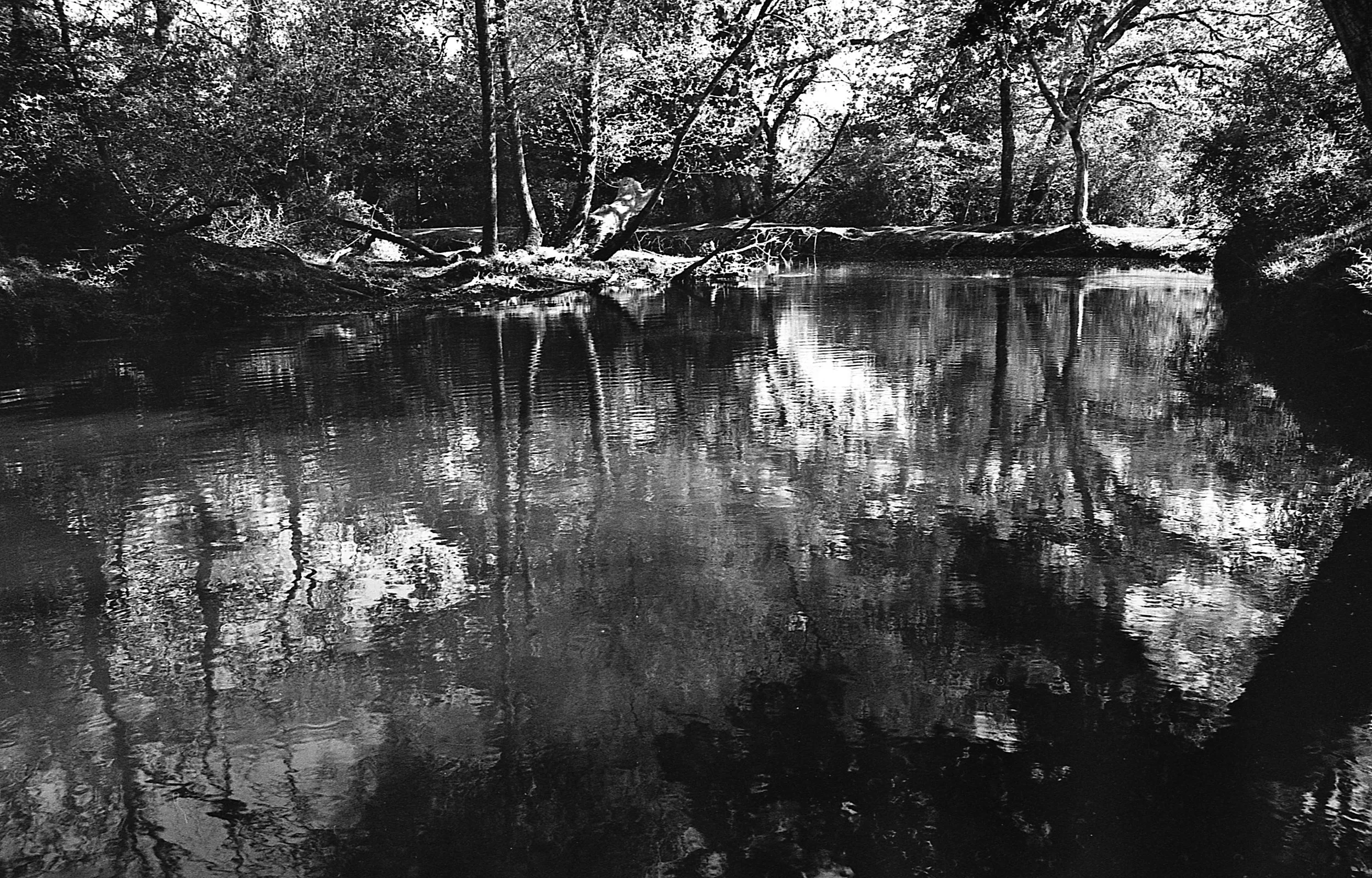 A black and white photograph of a calm river flowing through a wooded area with trees on both sides, reflections of the trees visible in the water.