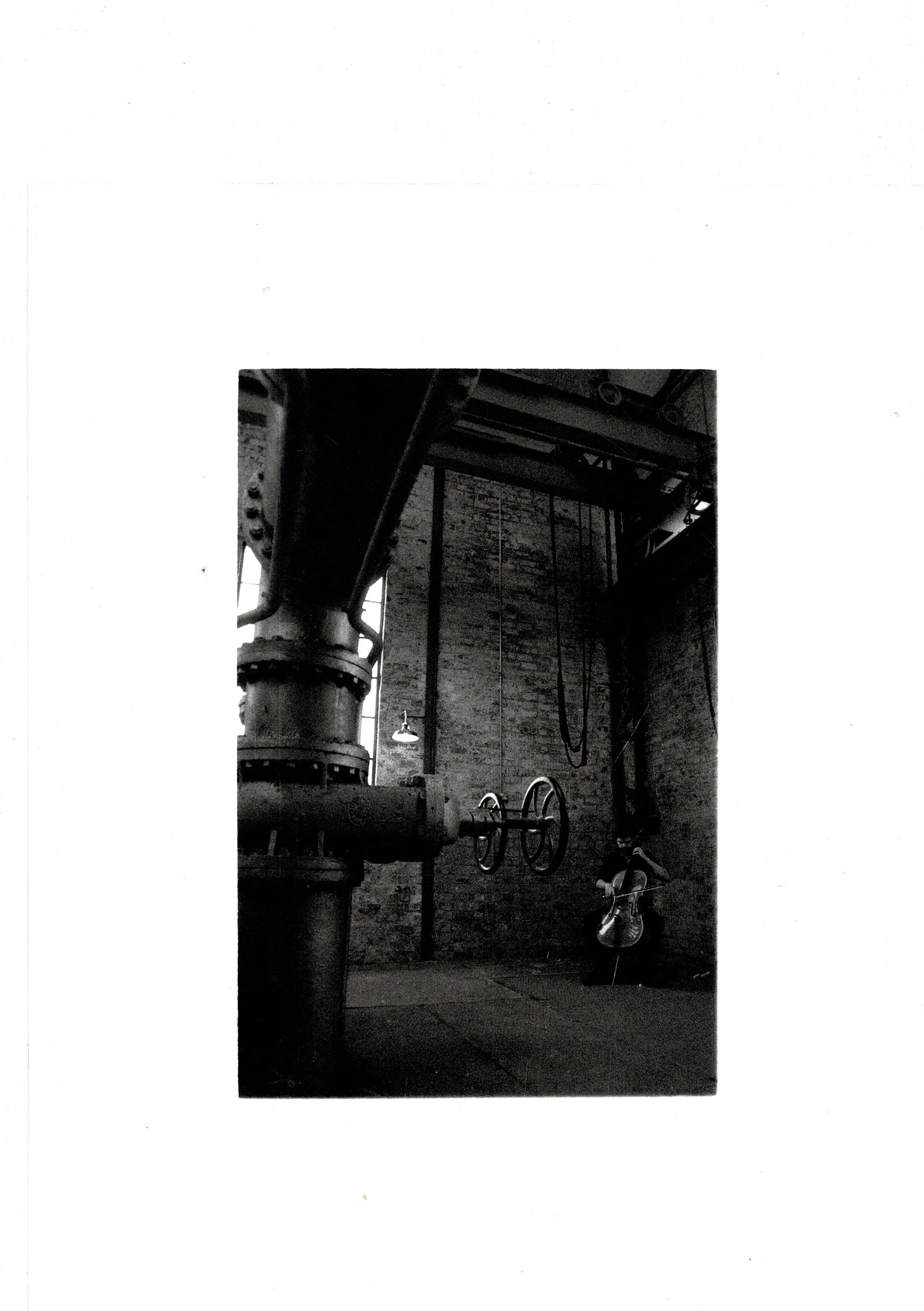 Black and white photograph of a cello standing among industrial pipework inside Scapa Flow Museum, hand-printed darkroom analogue print