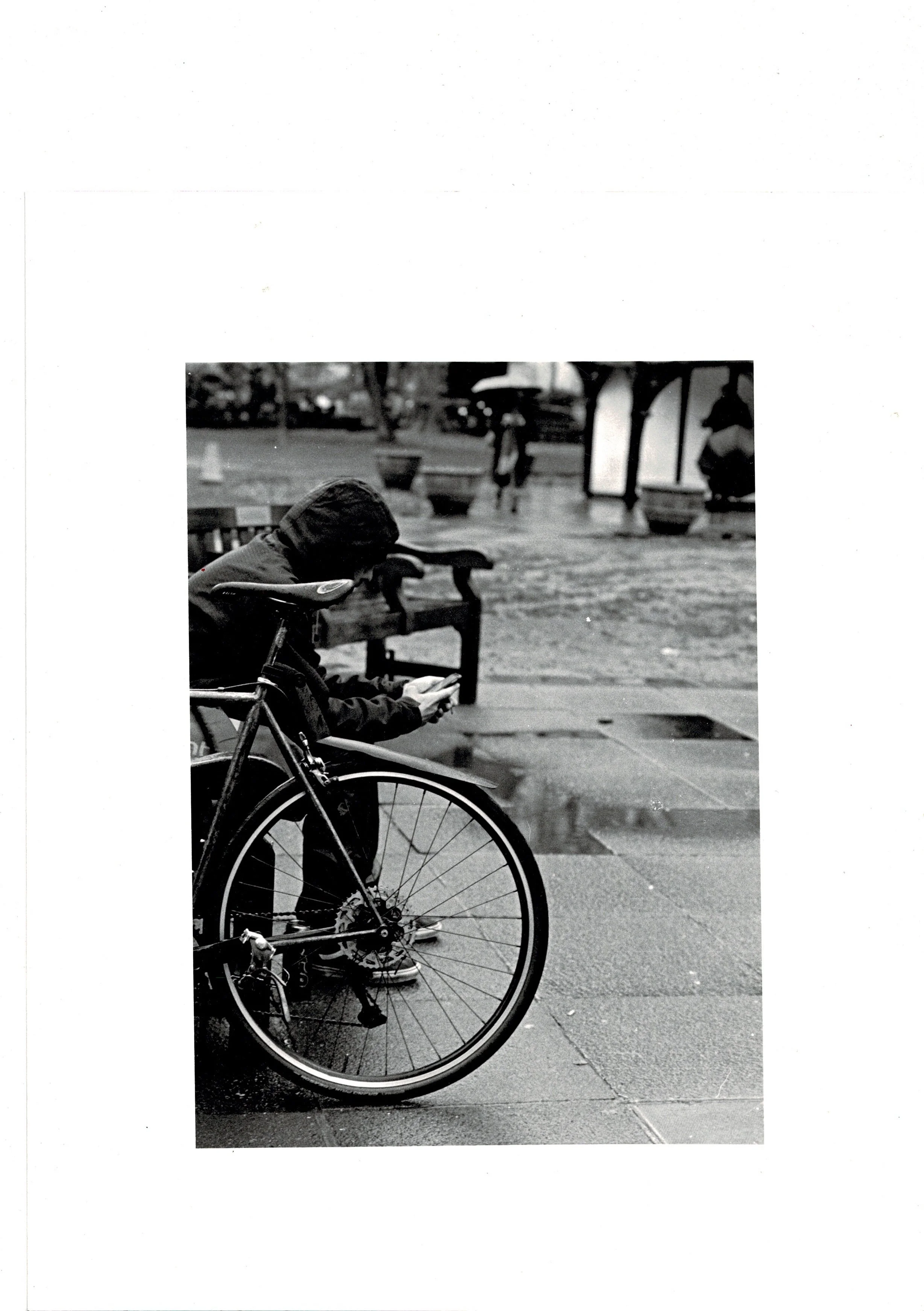 Black and white photograph of a cycle messenger leaning on railings beside his bike in Soho Square on a rainy day, hand-printed darkroom analogue print