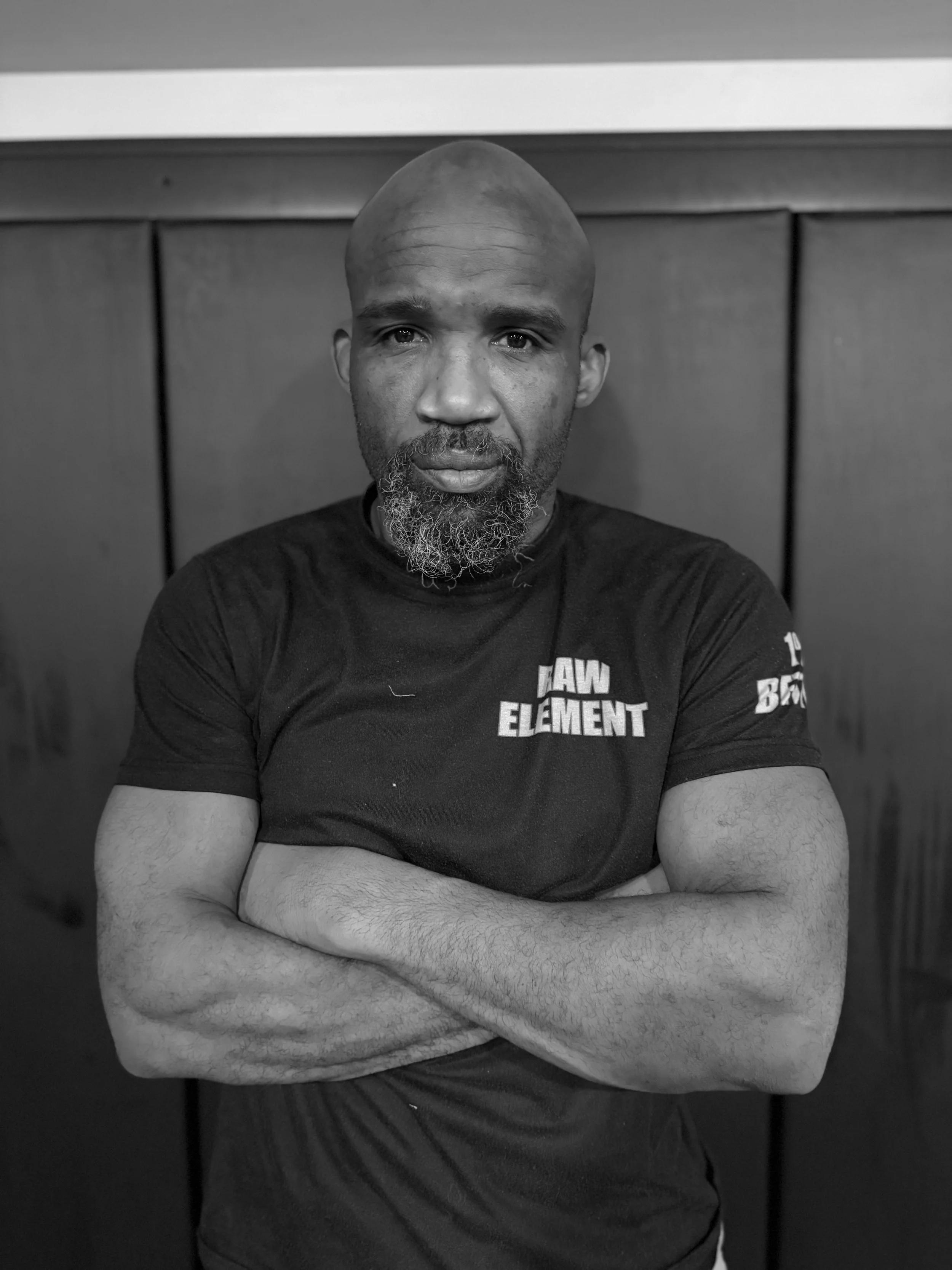 Black and white photo of a man with a shaved head and beard, crossing his arms and looking at the camera, wearing a T-shirt with 'RAW ELEMENT' written on it, standing against wooden paneling.