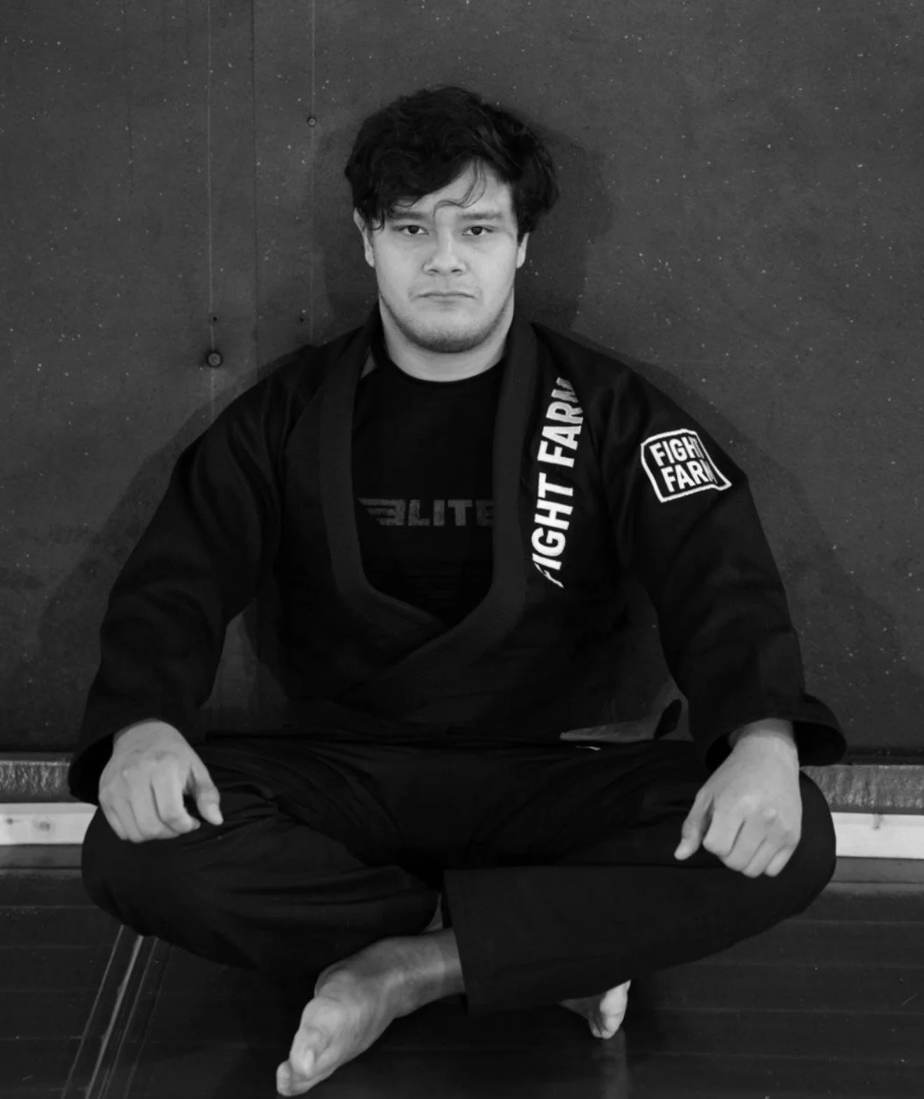 A young man sitting cross-legged on the floor in a martial arts gi, with a serious expression, against a dark wall.