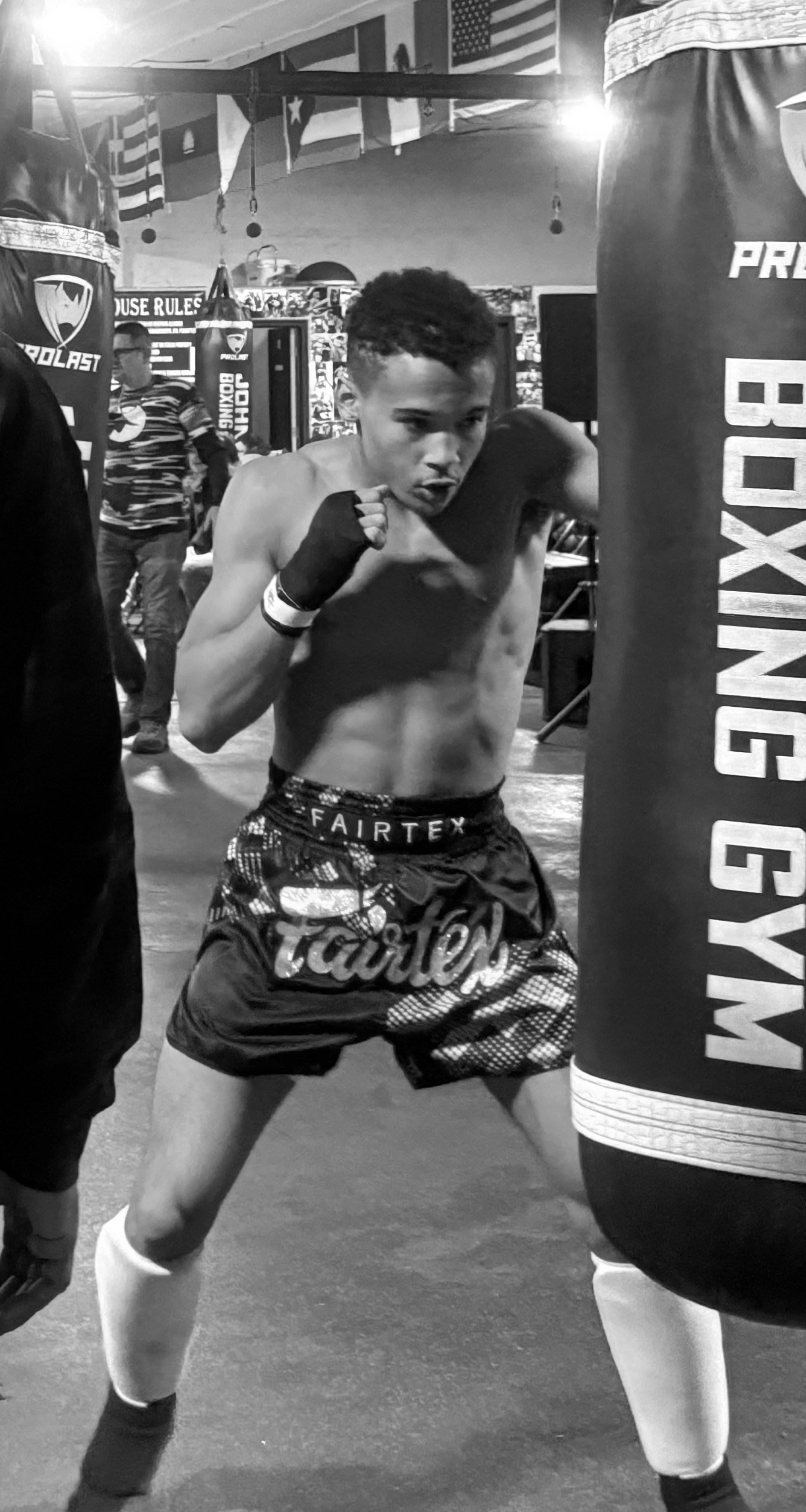 A young male boxer training in a gym, wearing boxing shorts, hand wraps, and boxing shoes, practicing punches near a punching bag.