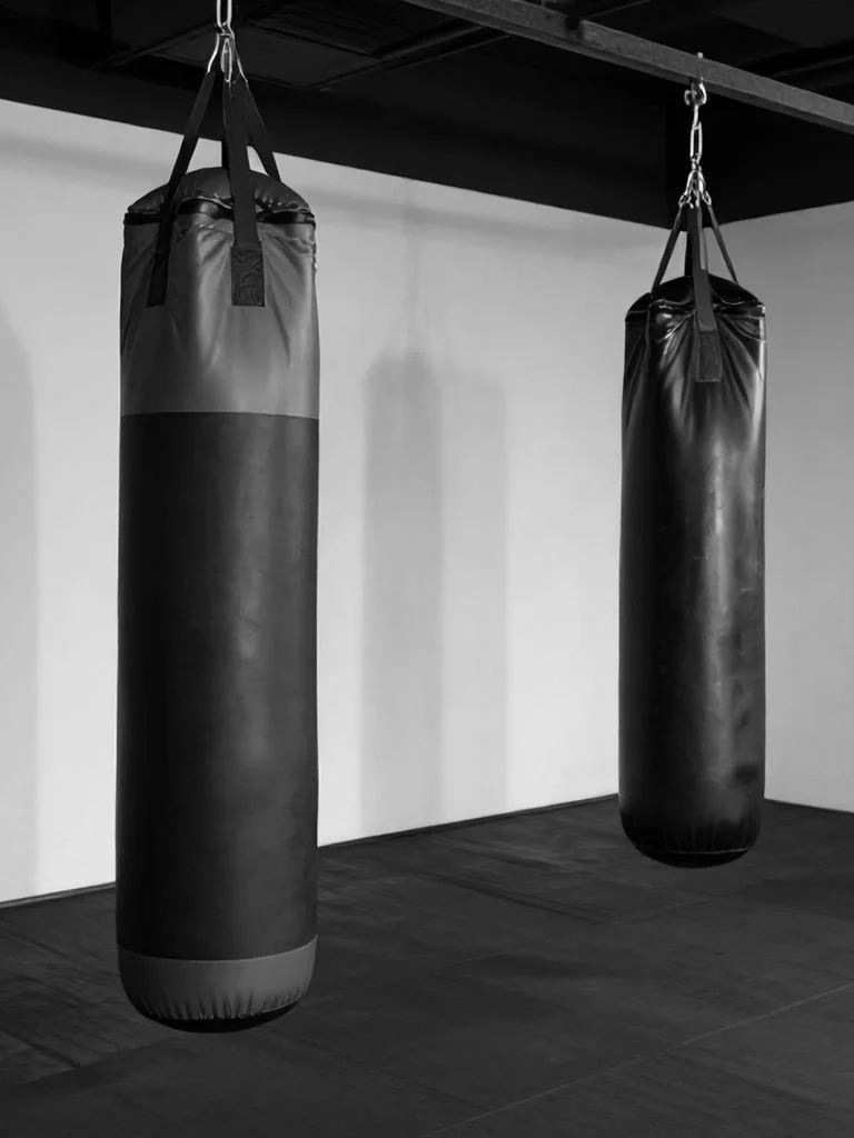 Two heavy punching bags hanging from a ceiling in a gym with wooden floors.