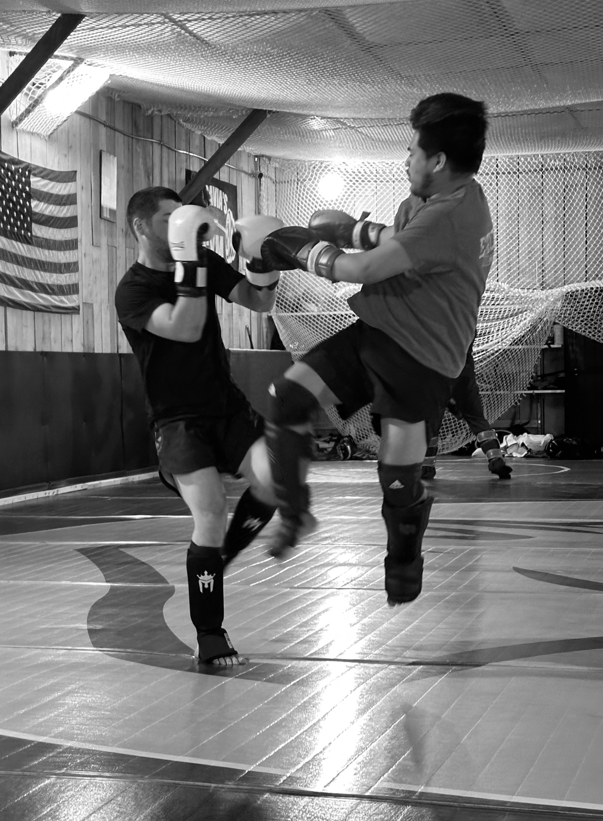 Two men are practicing martial arts in a gym, with one throwing a punch and the other in a defensive stance. The gym has wooden walls, a netted ceiling, and an American flag hanging on the wall.