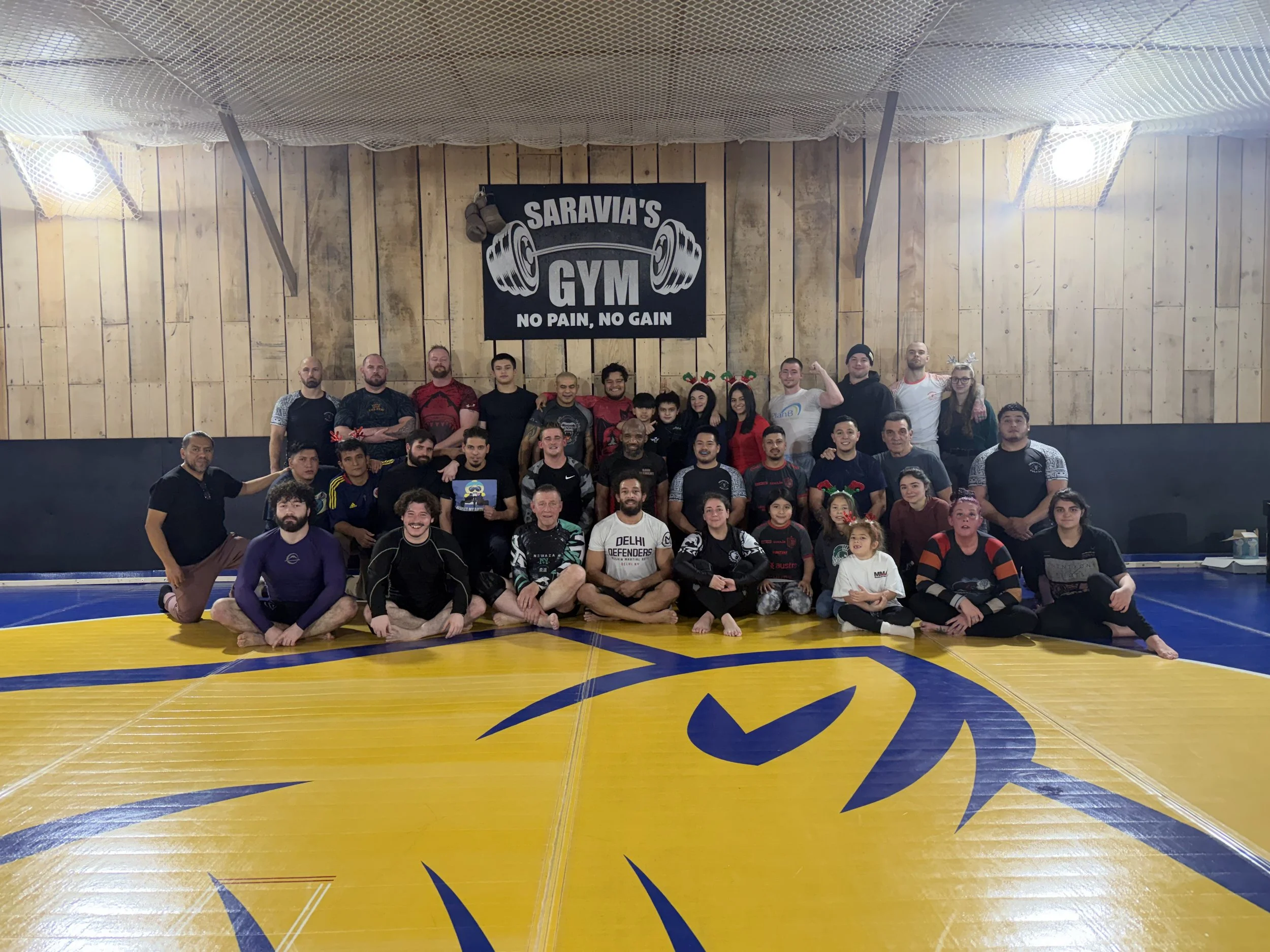 Group of people posing on a gym mat inside a martial arts gym with a wooden wall background and a sign that reads 'Saravia's Gym No Pain No Gain'.