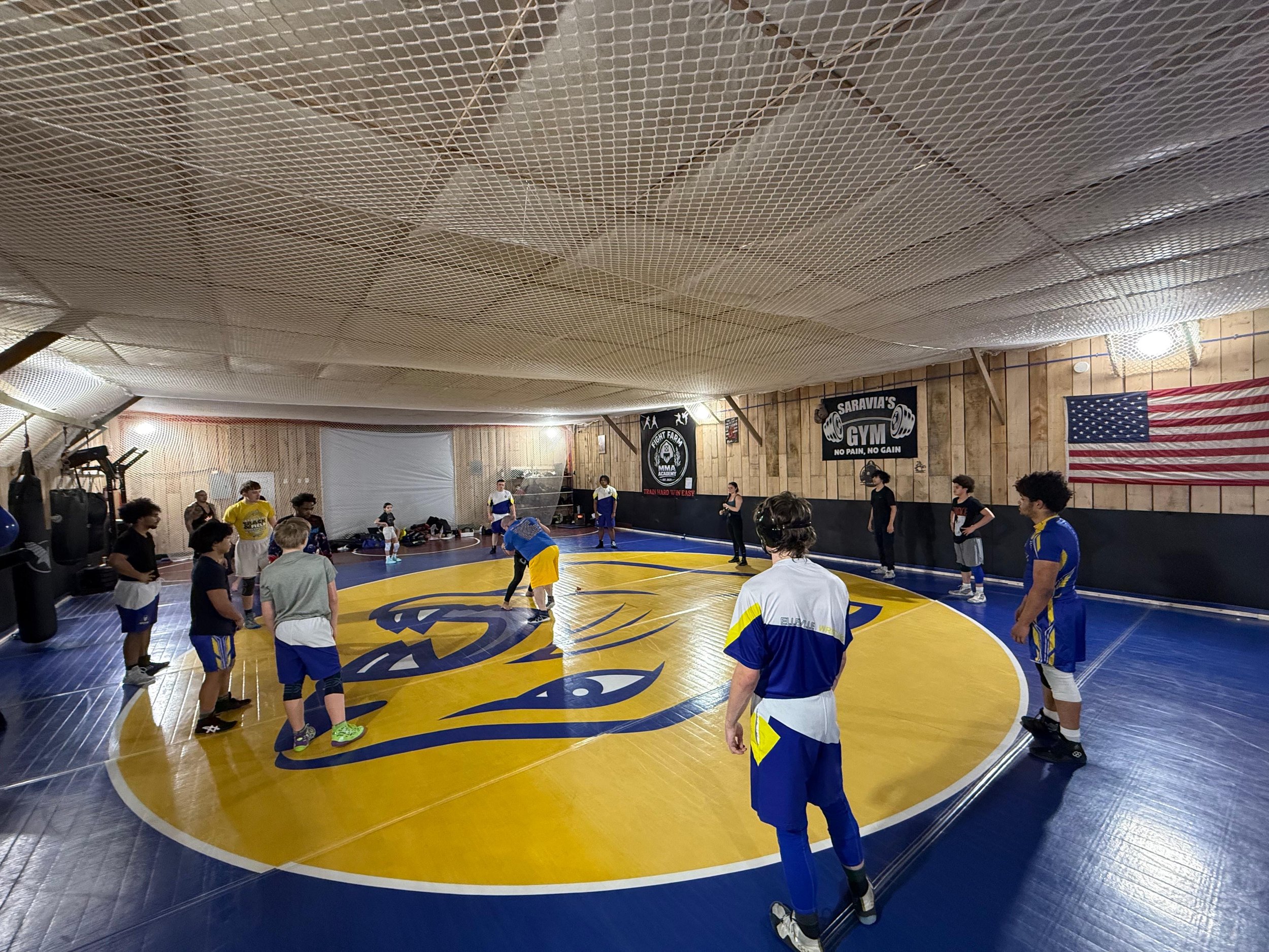 Boxing or wrestling gym with a group of young people standing on a circular mat with a logo, arranged in a circle. The gym has wooden walls with sports banners, including an American flag, and a large netted ceiling. Some equipment is visible along the sides.