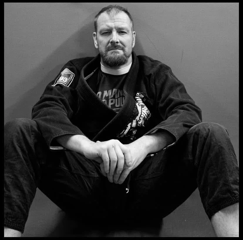 A man with a beard sitting on the floor with his legs spread apart, wearing a martial arts gi and a T-shirt underneath, against a plain wall.