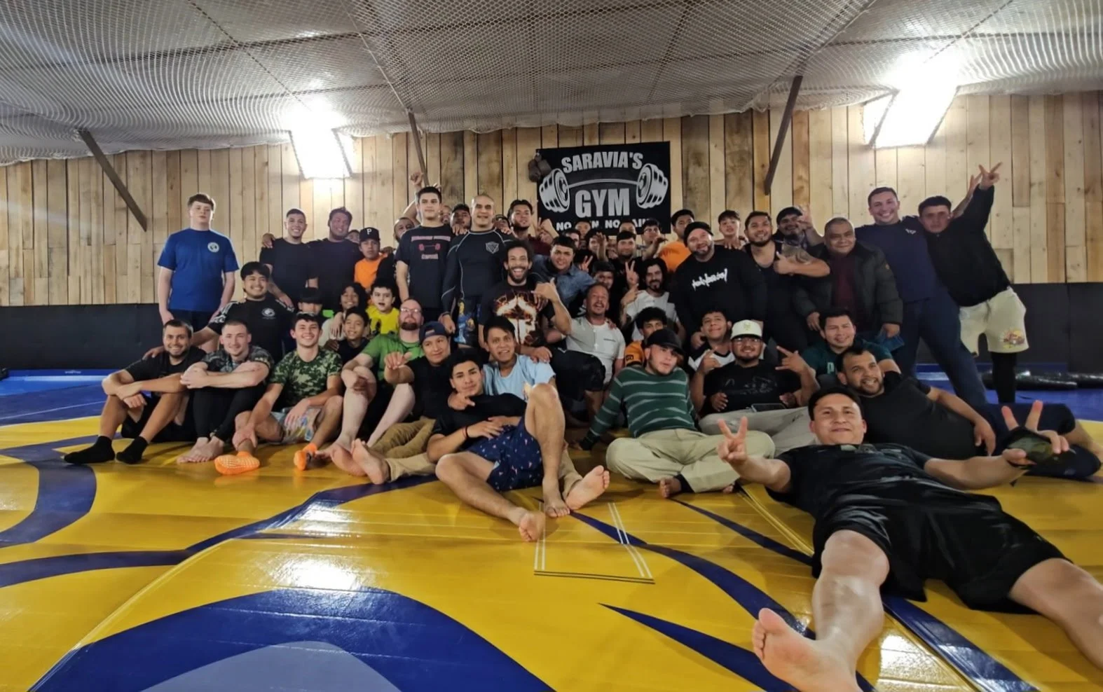A group of men and children gathered together in a gym, posing for a group photo on a colorful wrestling mat. Everyone is smiling and some are making peace signs or victory signs. The background features a sign that says 'SaraVias Gym' with weightlifting symbols.